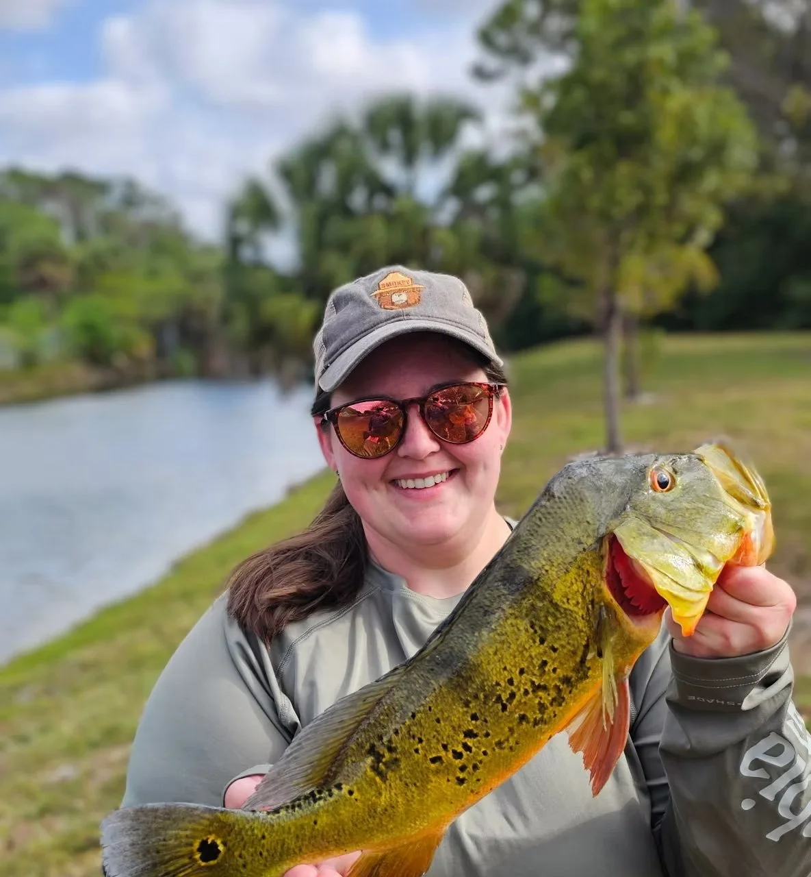 A woman wearing sunglasses and a cap, smiling while holding a large fish near a riverbank with green trees and cloudy sky in the background.