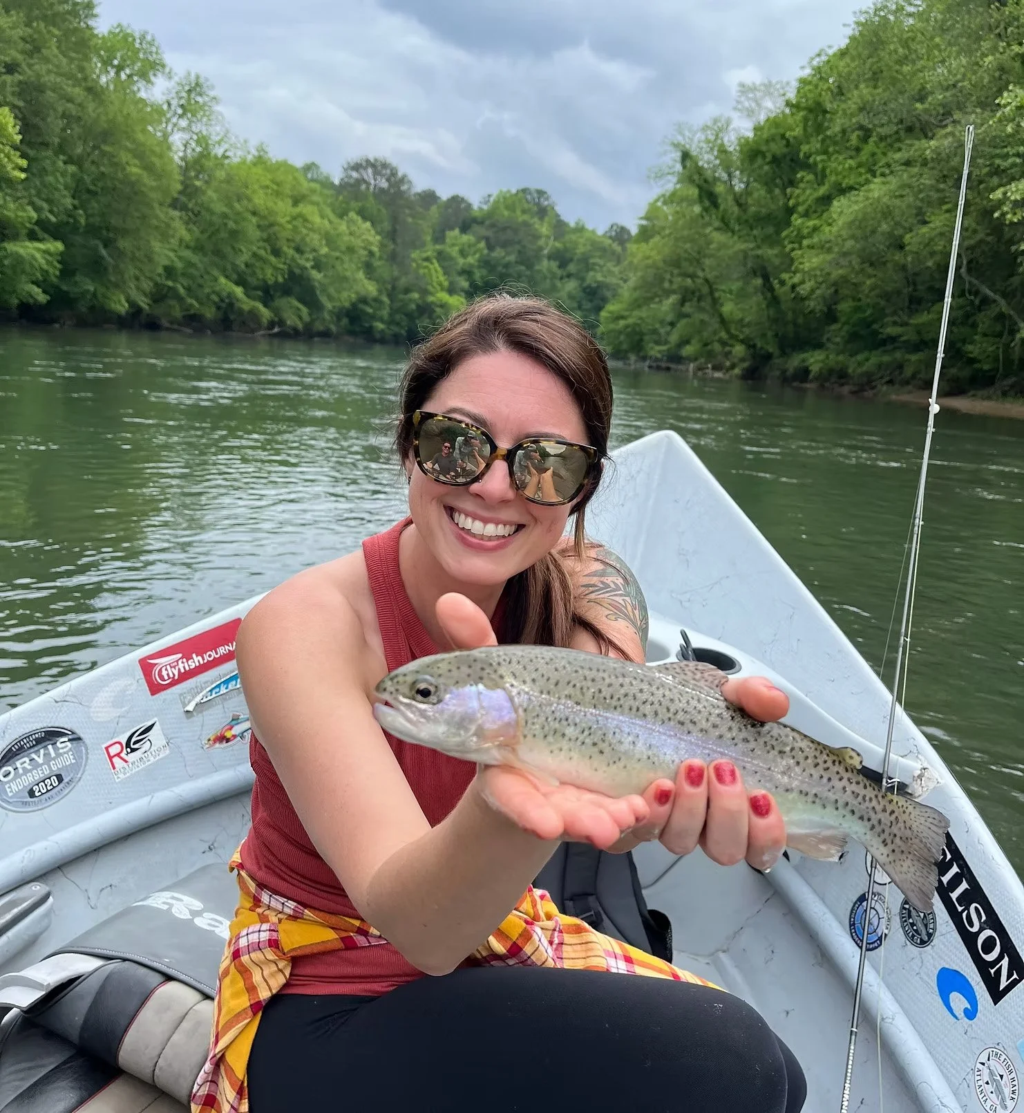 A smiling woman wearing sunglasses on a boat, holding a fish with green and purple scales, surrounded by a river and green trees.
