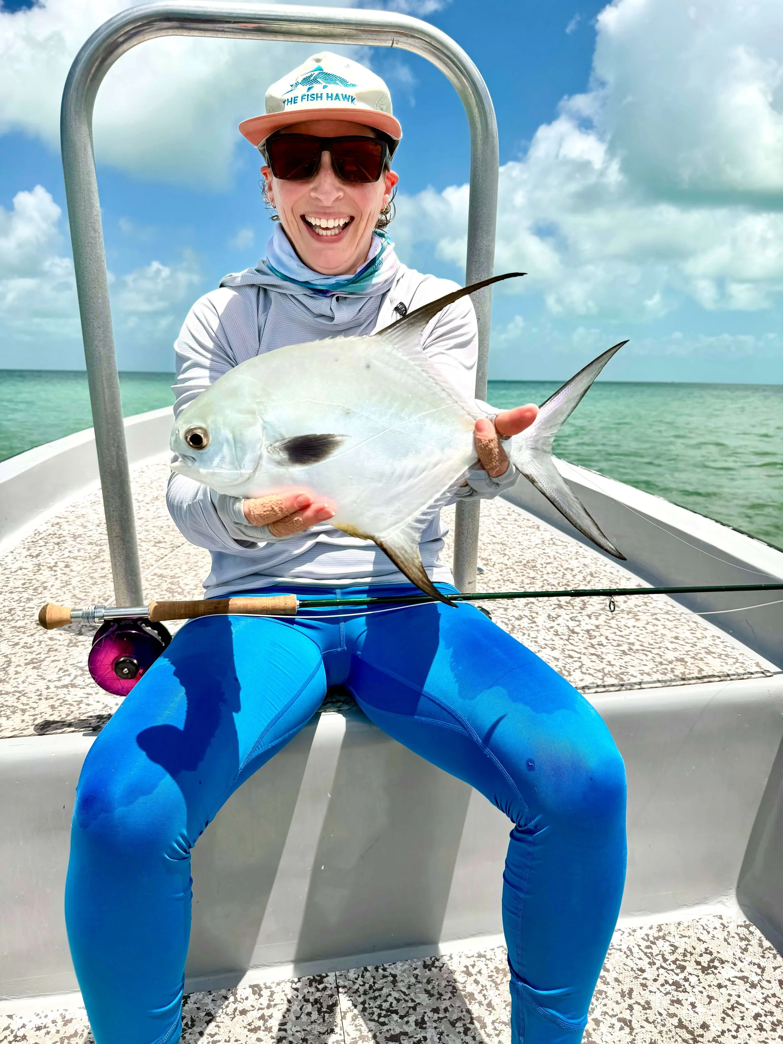 Person sitting on a boat holding a fish, wearing sunglasses, a cap, and bright blue pants with a fishing rod on their lap, against a background of blue sky and ocean.