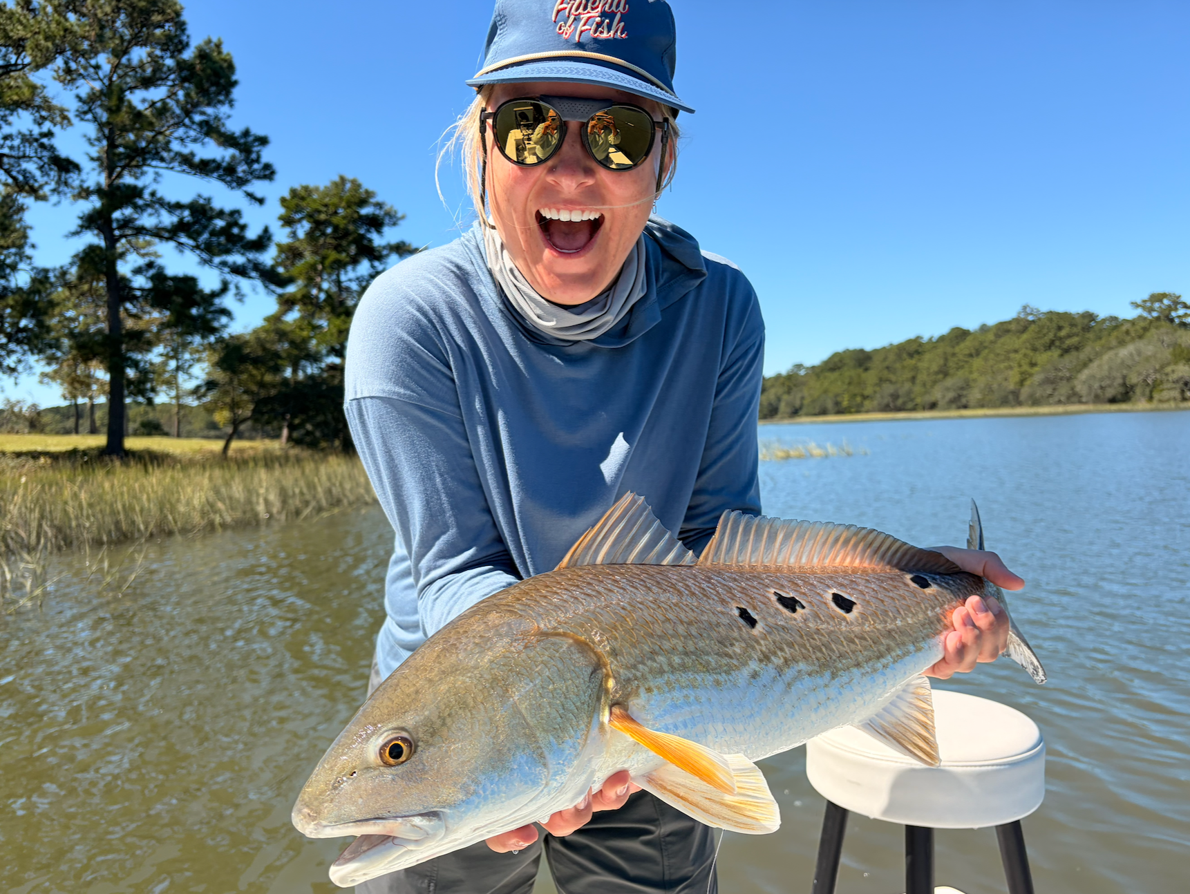 Woman smiling and holding a large colorful fish by a river with green trees and water in the background.