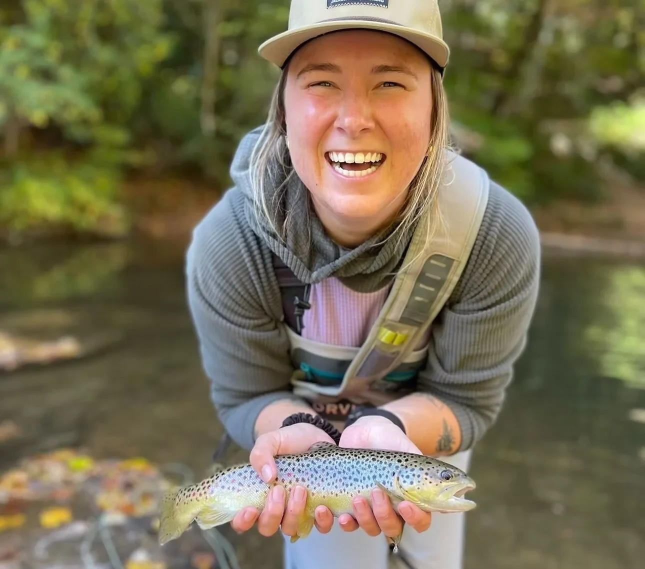 Woman smiling and holding a small colorful fish by a river with green trees in the background.