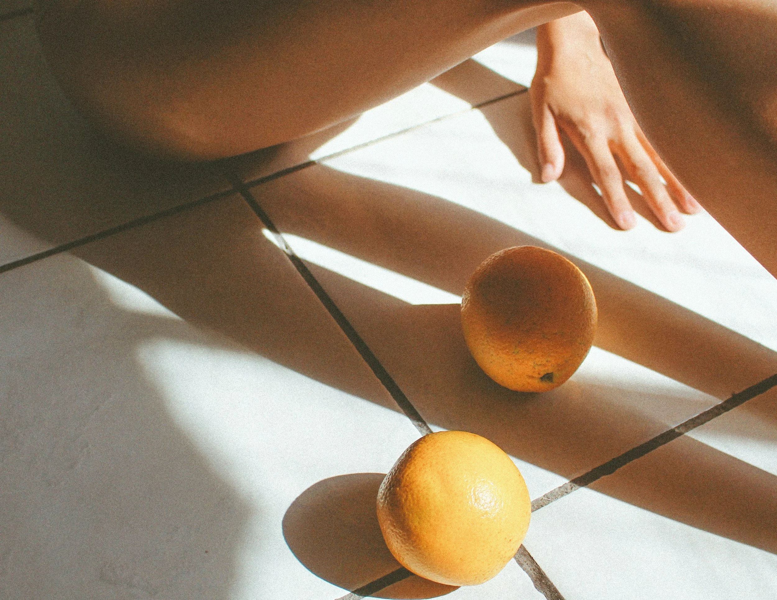 A person sitting on a tiled floor with their legs extended, two oranges are placed on the floor, and the person's hand is reaching towards their foot.