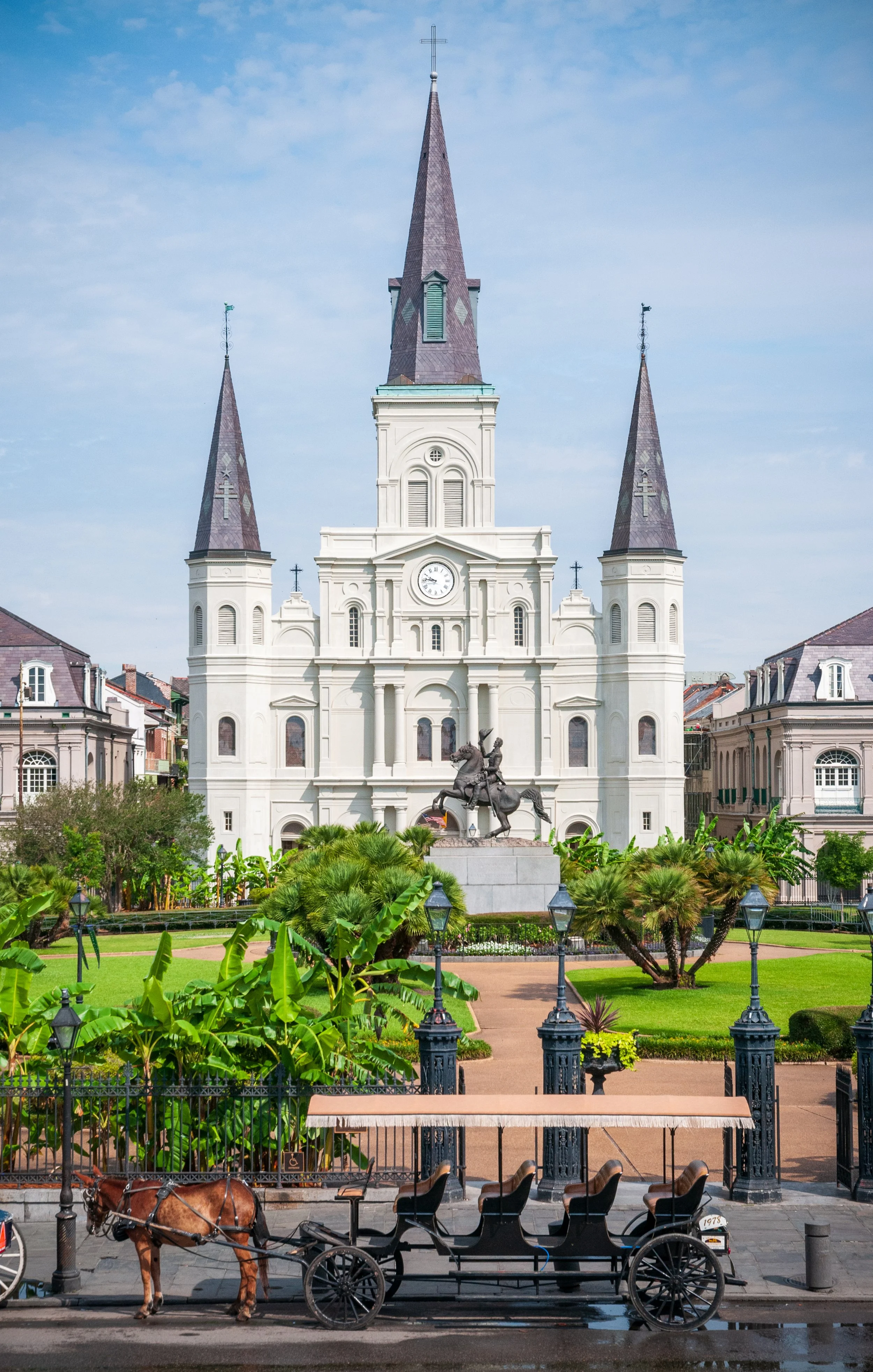 A white church with three tall spires and a clock, surrounded by green parks, trees, and a horse-drawn carriage in the foreground.