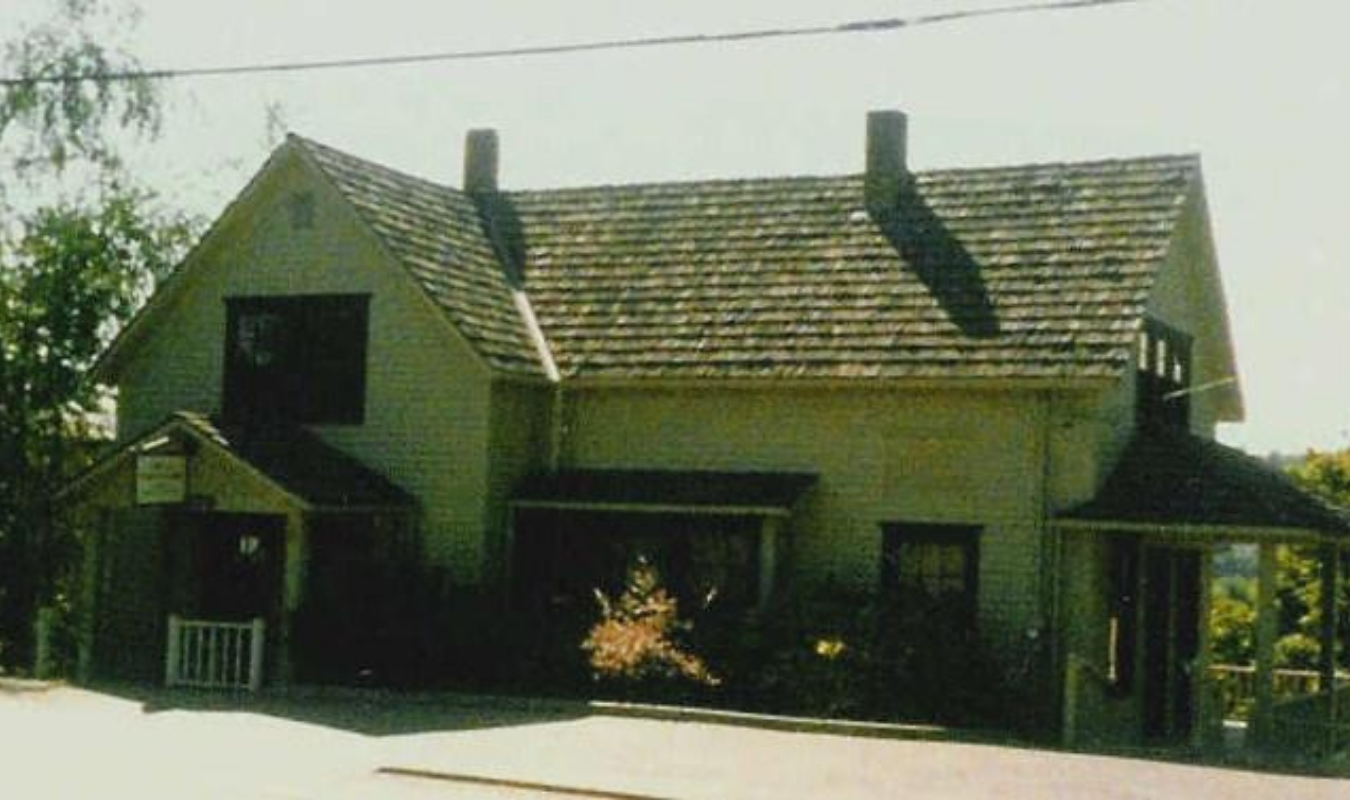 A two-story house with a gable roof, chimney, and porch, surrounded by trees and shrubbery.