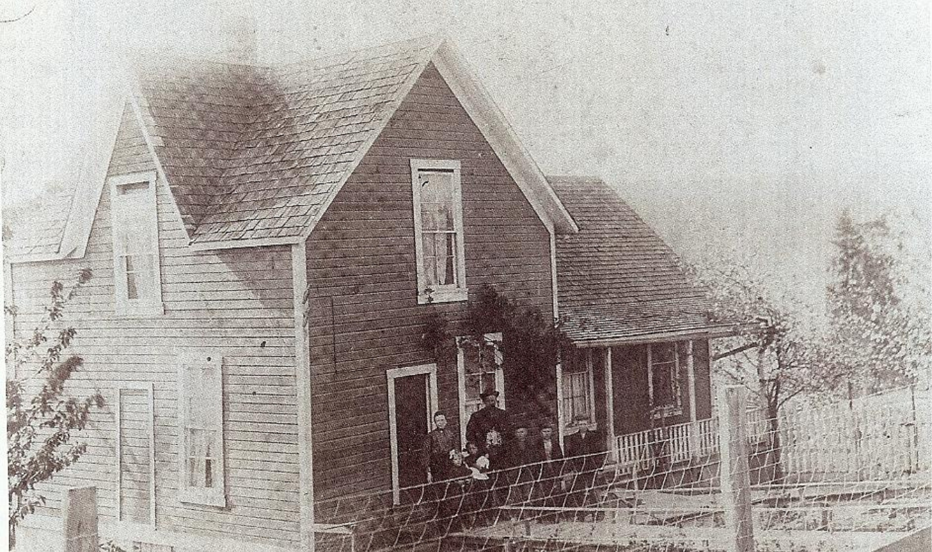 A black and white photo of an old wooden house with a porch, located in a rural area. There are five people standing on the porch in front of the house, with a wooden fence surrounding the property. The house has a steep gabled roof and tall narrow windows.