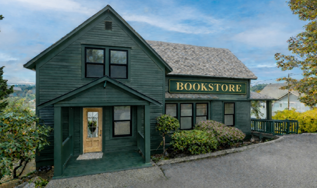A green, two-story building with a sign that reads "BOOKSTORE" on the front. The building has several windows, a small front porch with a wooden door, and is surrounded by bushes and a driveway.