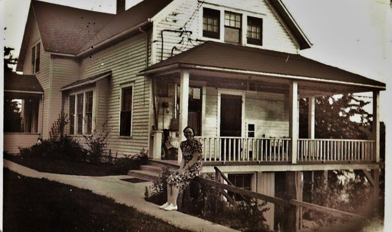 A woman standing on a sidewalk in front of a two-story house with a porch, white siding, and multiple windows, holding a coffee cup and smiling.