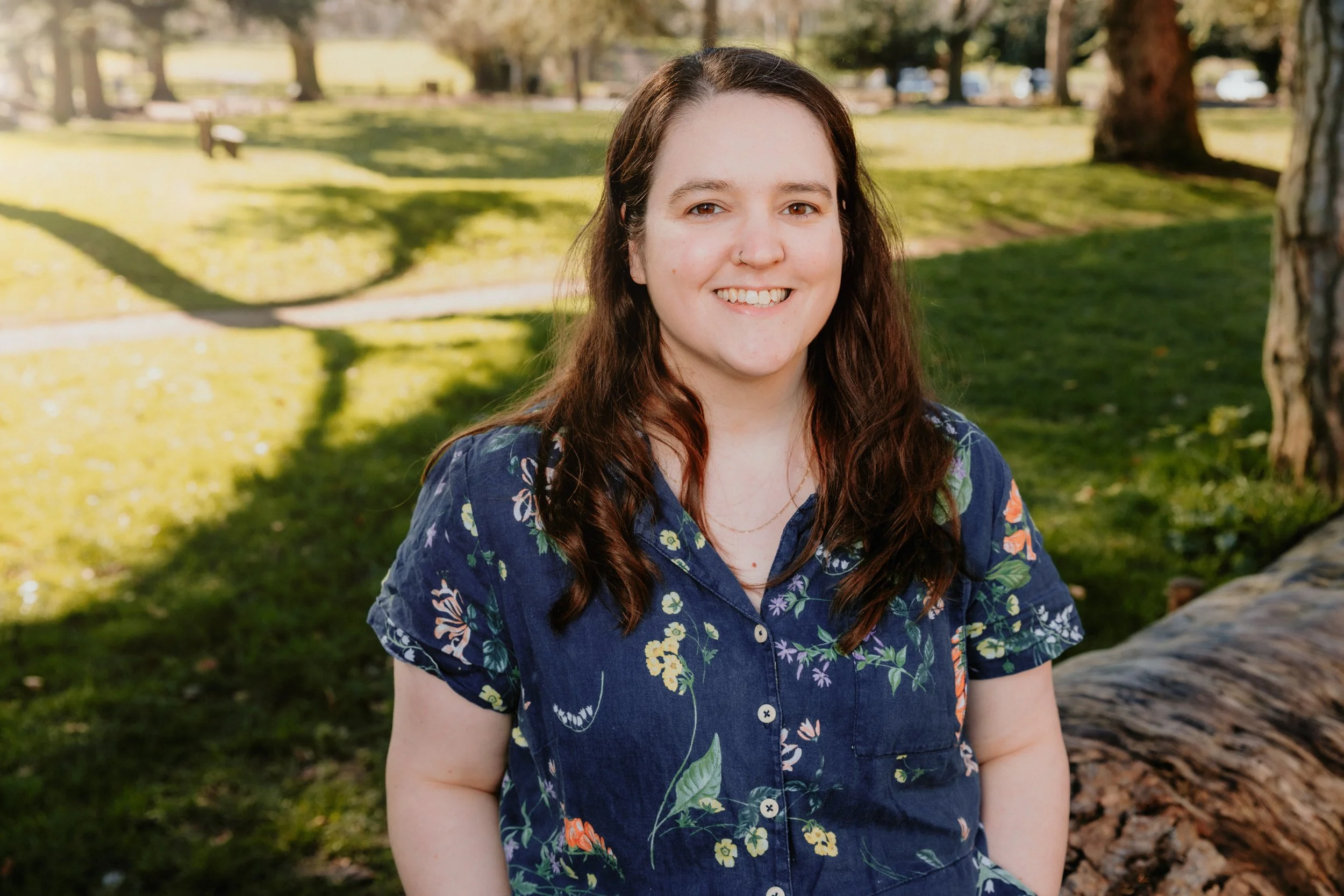 A young woman with long brown hair, wearing a floral patterned blue shirt, smiling, outdoors in a park with grass and trees in the background.