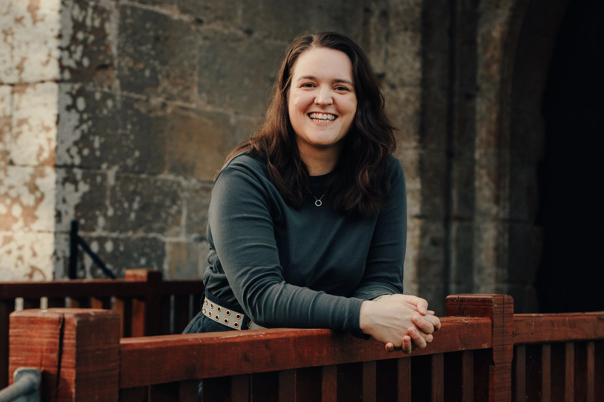 A young woman with long dark hair and a big smile, wearing a dark long-sleeve shirt and a silver belt with eyelets, leaning on a wooden railing in an outdoor setting with a brick wall background.