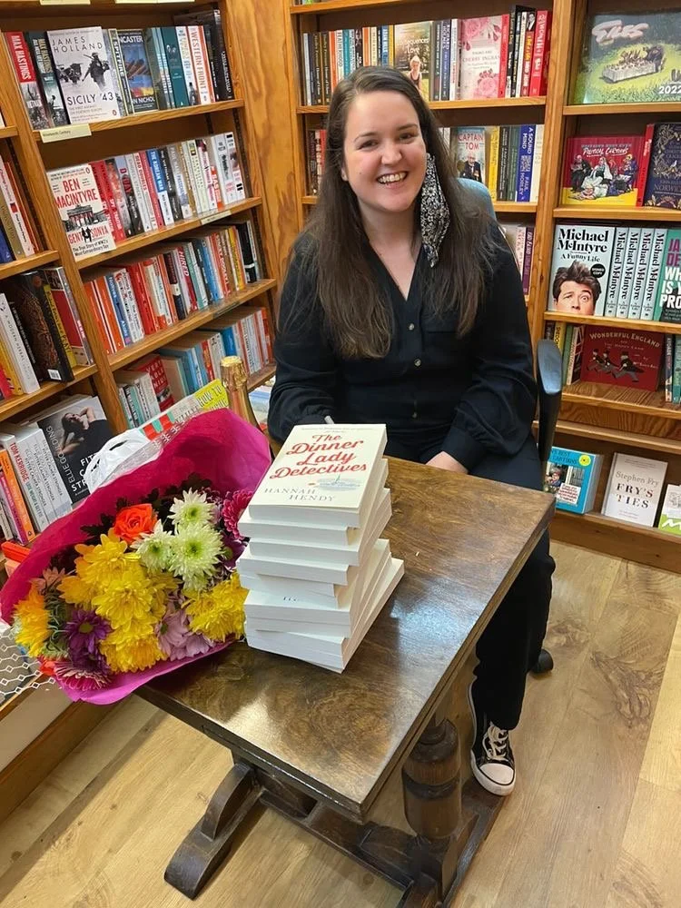 Young woman with long brown hair sitting at a book signing event, smiling, with a bouquet of flowers and a stack of books titled 'The Dinner Lady Detectives' on the table, in a bookstore filled with shelves of books.