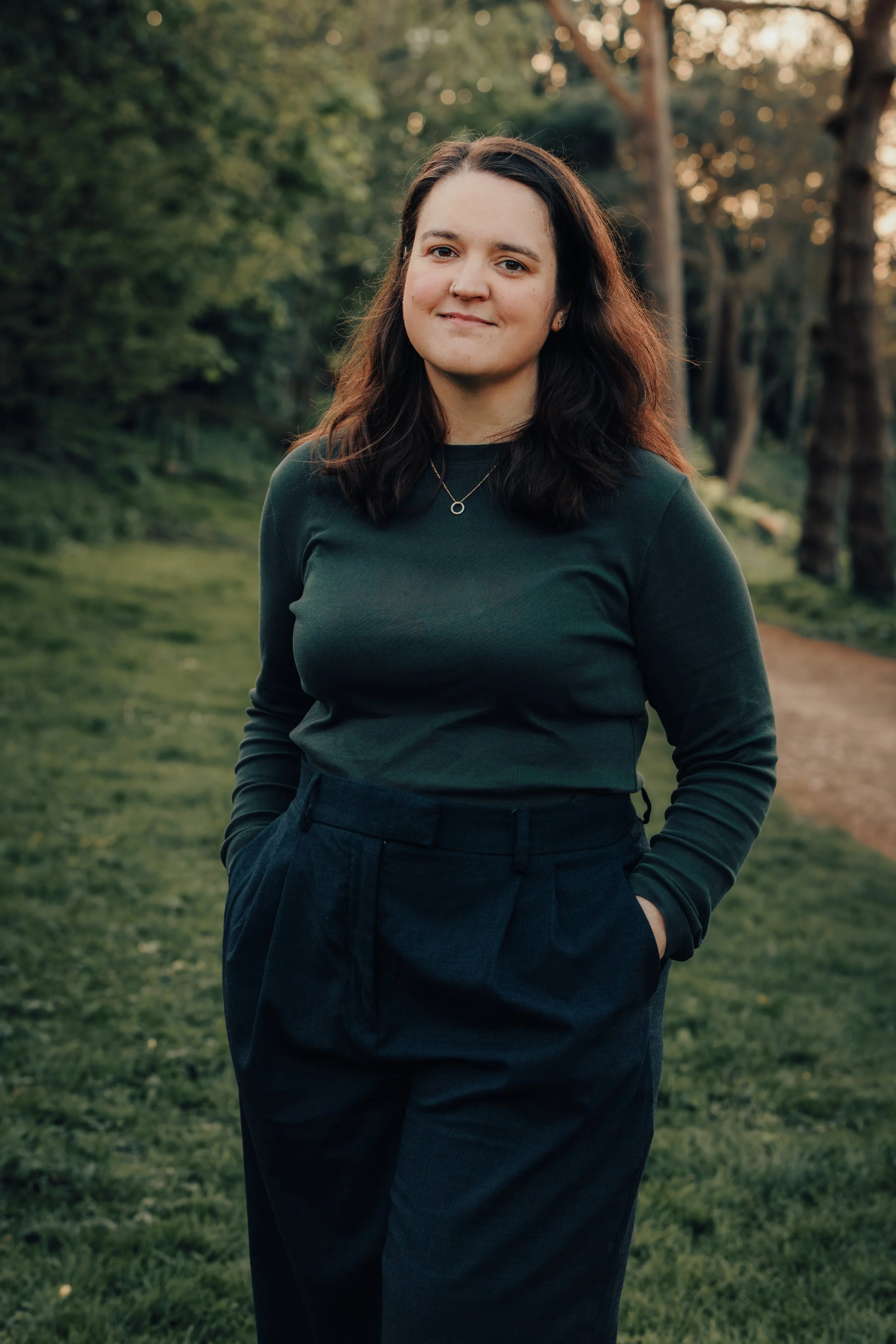A woman with dark hair standing outdoors in a park-like setting with trees and grass, wearing a dark long-sleeve top and black pants, smiling softly at the camera during what appears to be late afternoon or early evening.