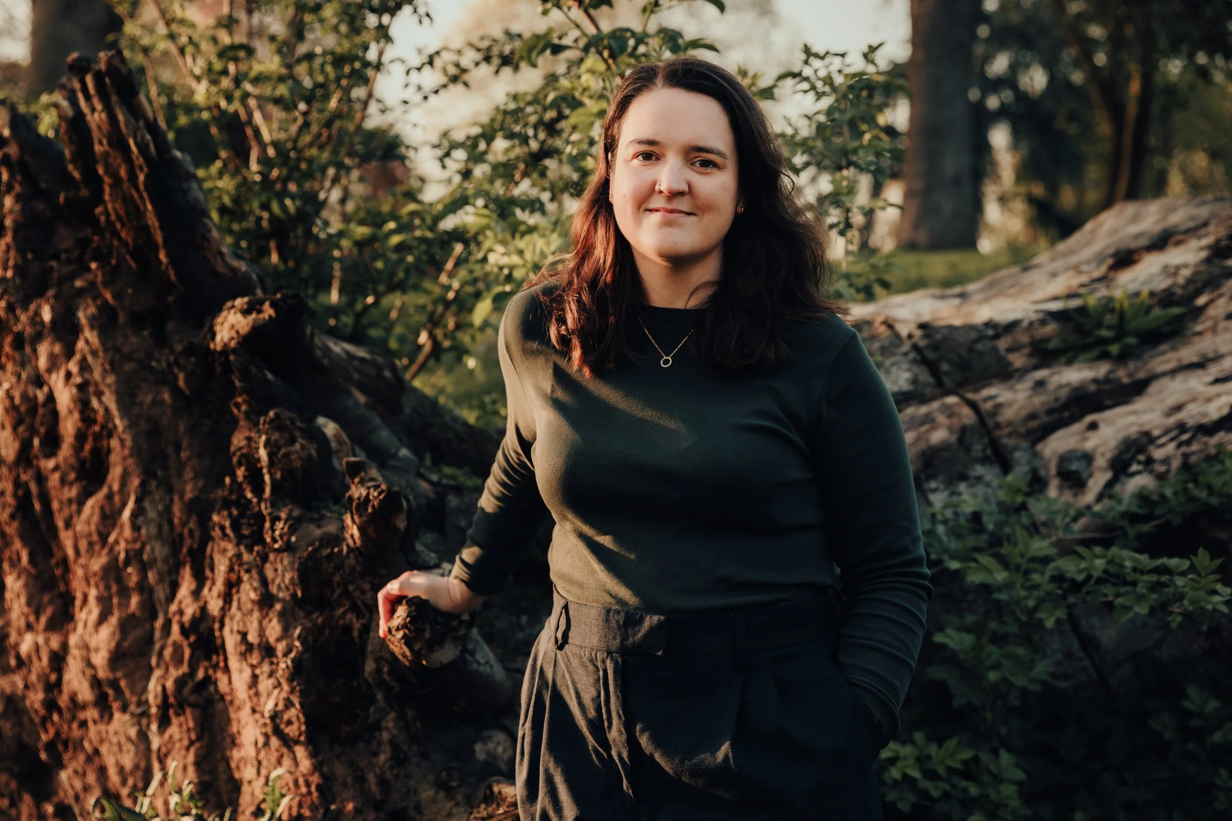 A young woman with dark brown hair, wearing a dark green long-sleeve shirt and black pants, is standing outdoors near a tree with fallen branches, in a park during golden hour.