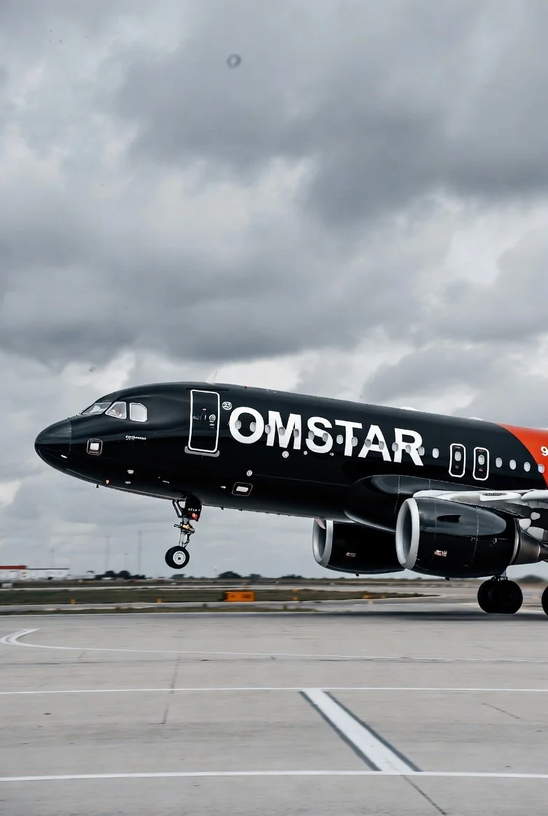A black OMSTAR airplane taking off from an airport runway on a cloudy day.