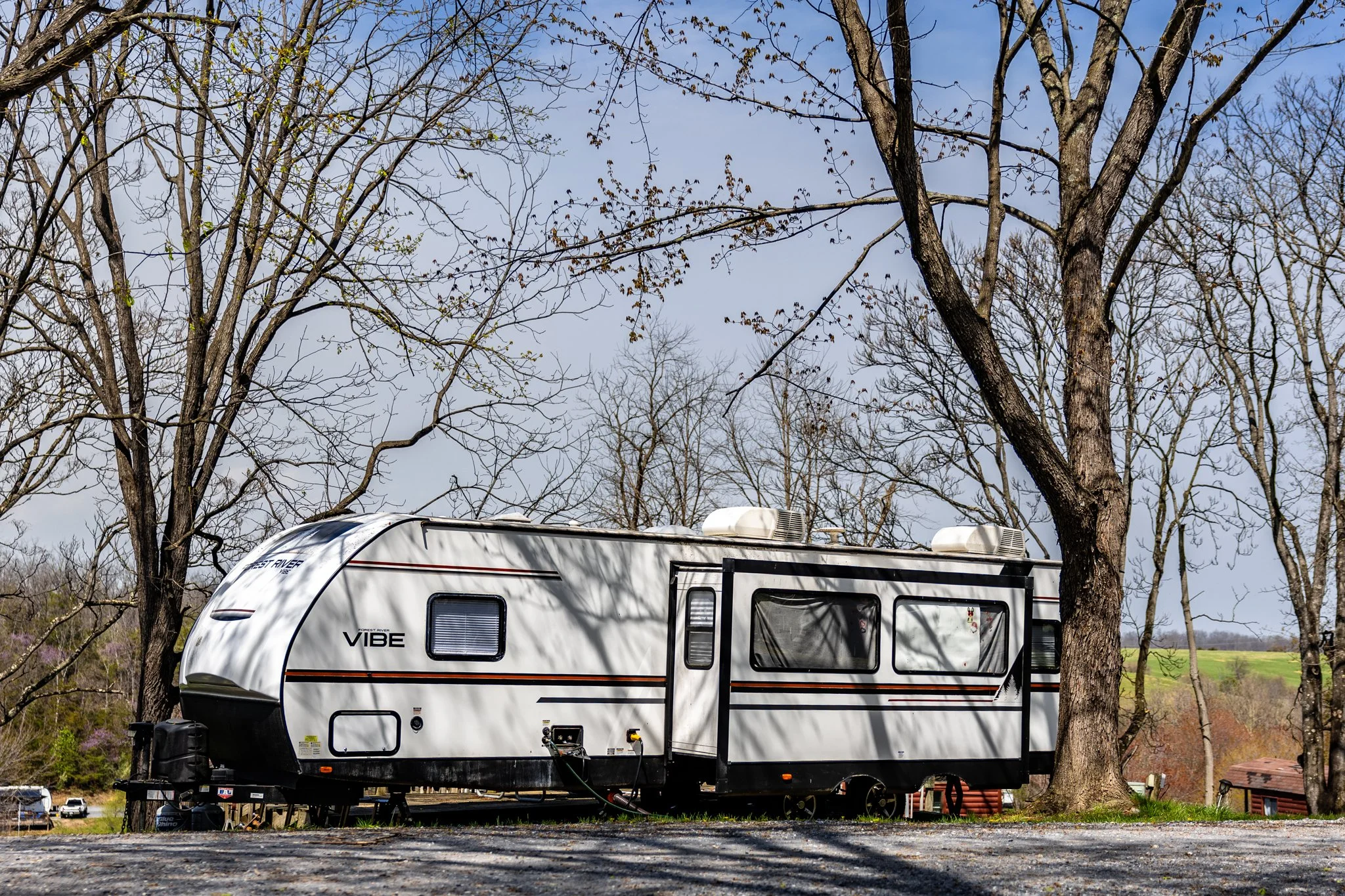 A white travel trailer parked in a wooded campsite under leafless trees on a sunny day.