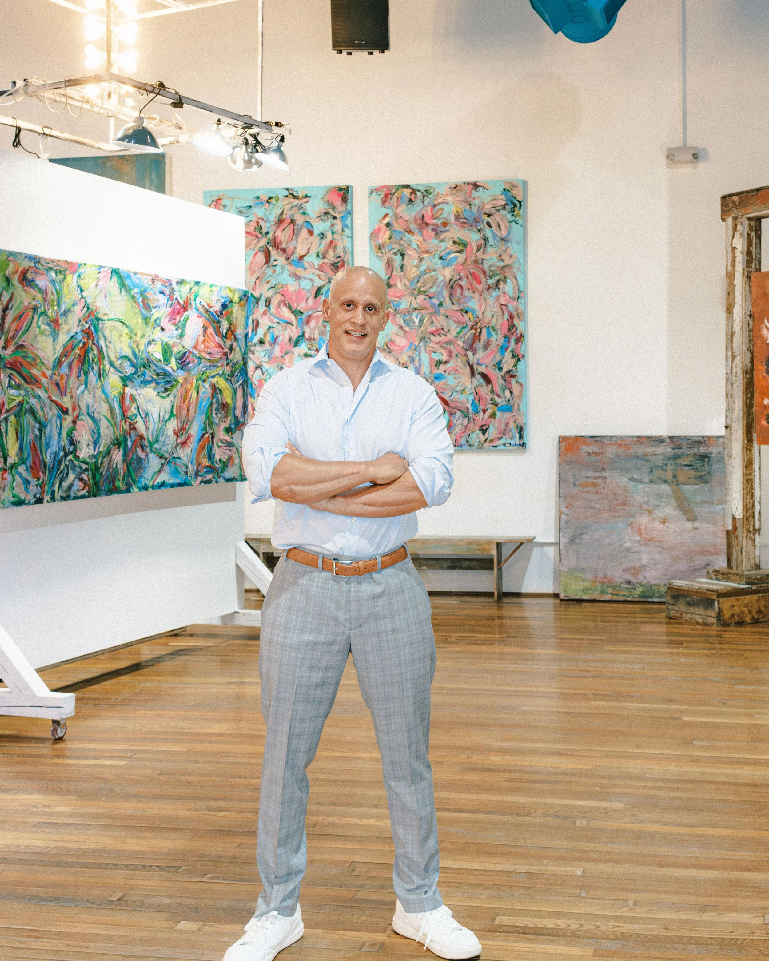 A man in a white shirt and gray plaid pants standing with arms crossed in an art gallery with colorful abstract paintings on the walls.