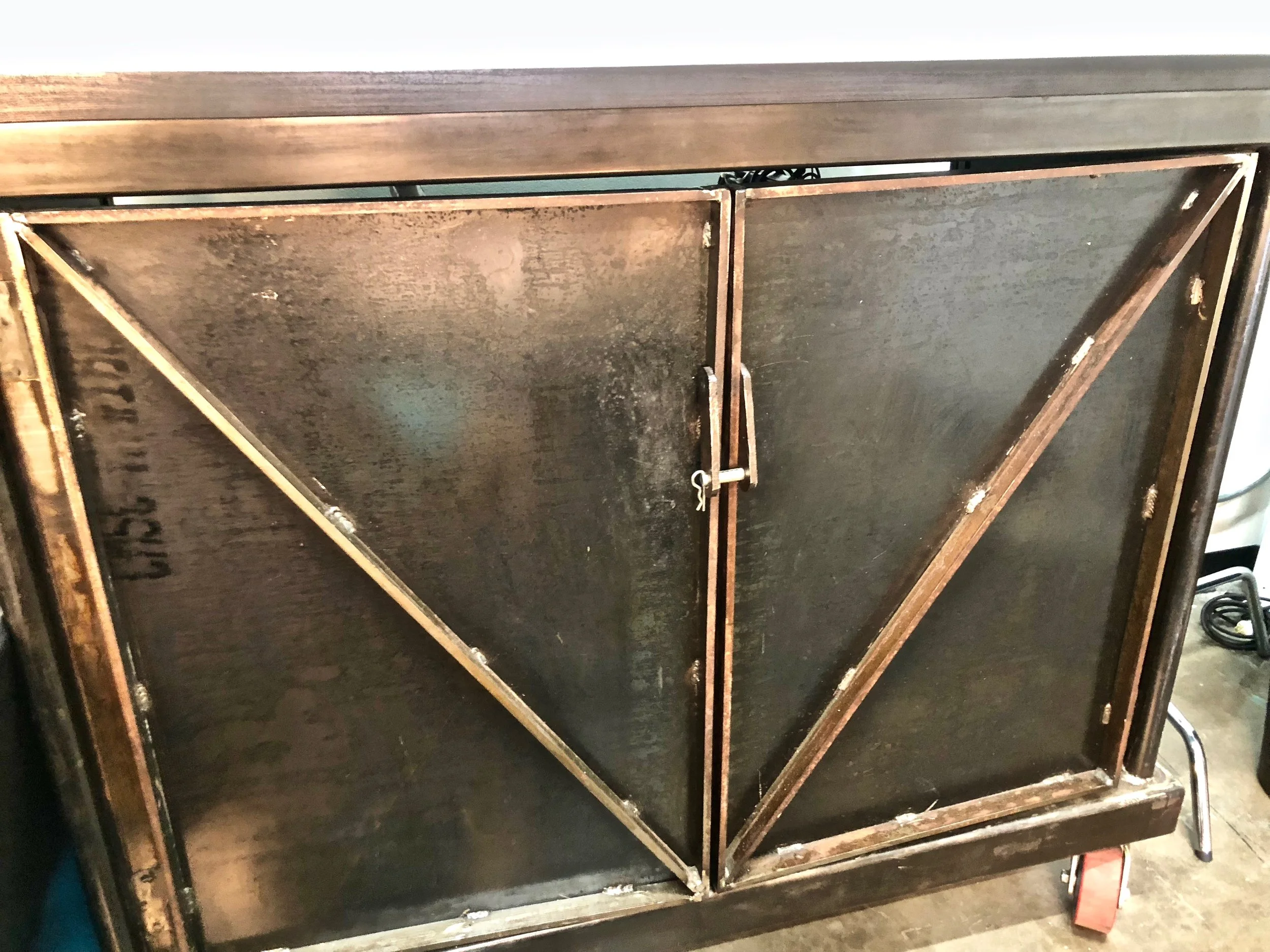 The image shows a front view of a black metal cabinet with two doors, secured by a small latch, with visible wear and rust. It is placed on a floor with some tools and equipment nearby.