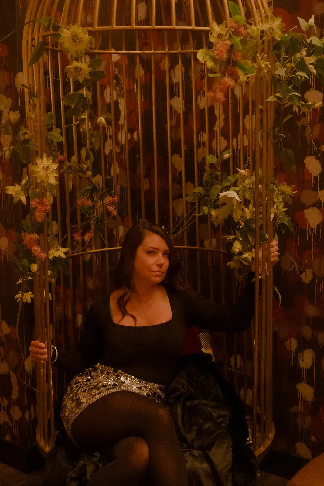 A woman sitting inside a decorative golden birdcage surrounded by floral arrangements and a patterned wallpaper background.