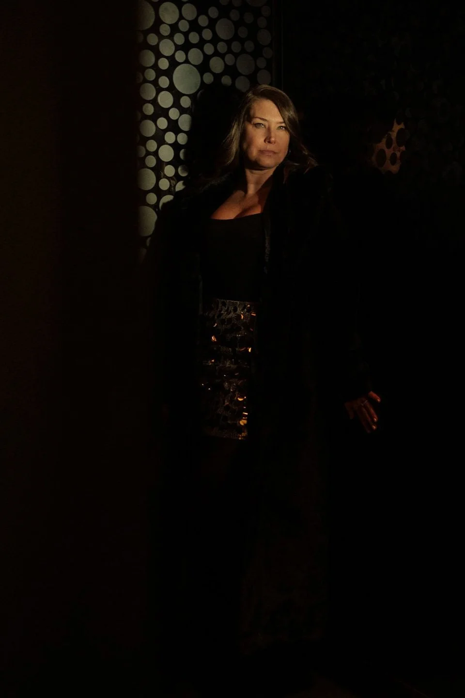 A woman standing in a dark environment, illuminated partially, with a patterned background featuring circles, dressed in a black outfit with a shiny, metallic skirt.