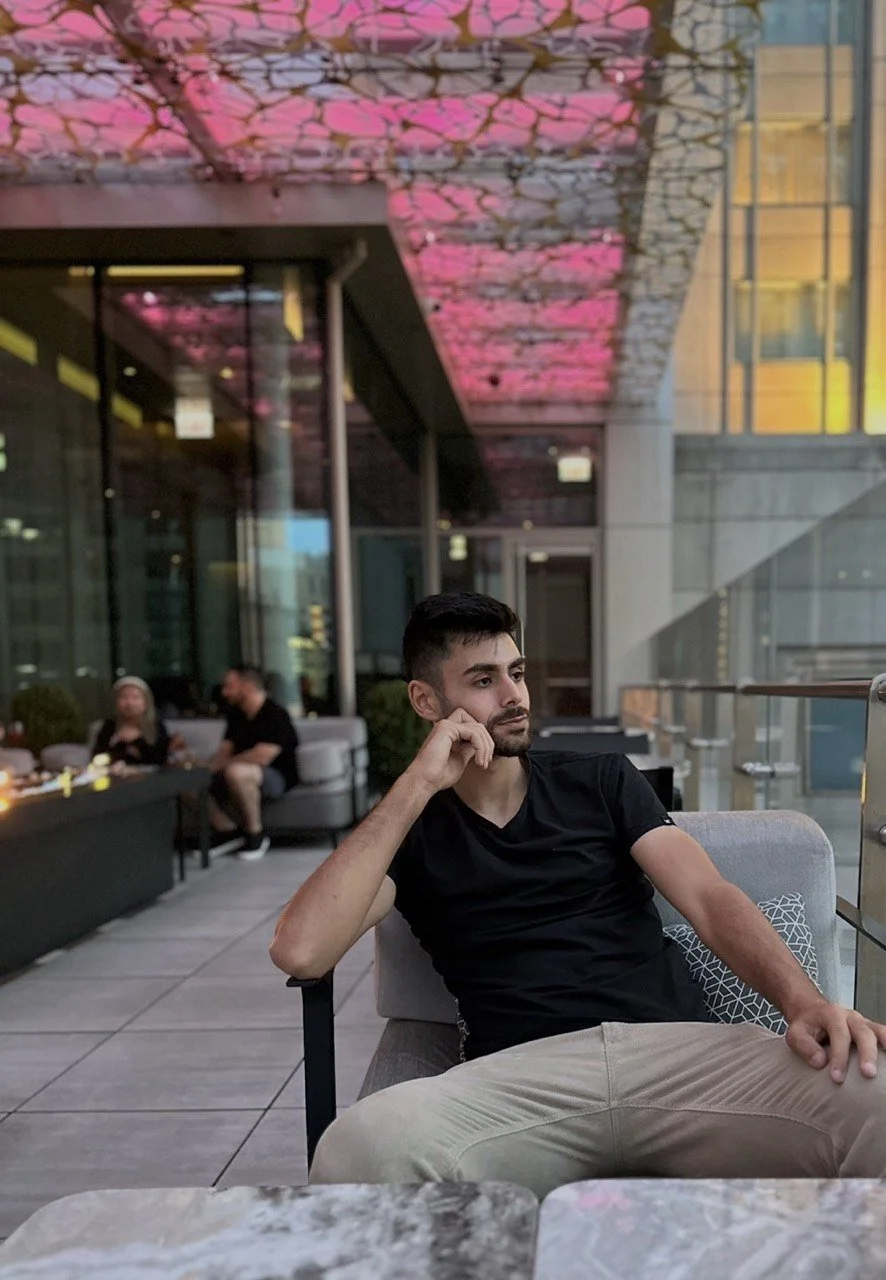 A man sitting on a chair at a modern outdoor cafe, with one hand resting on his knee and the other hand supporting his chin, looking thoughtfully to the side. There are other people seated in the background and colorful lights on the ceiling.