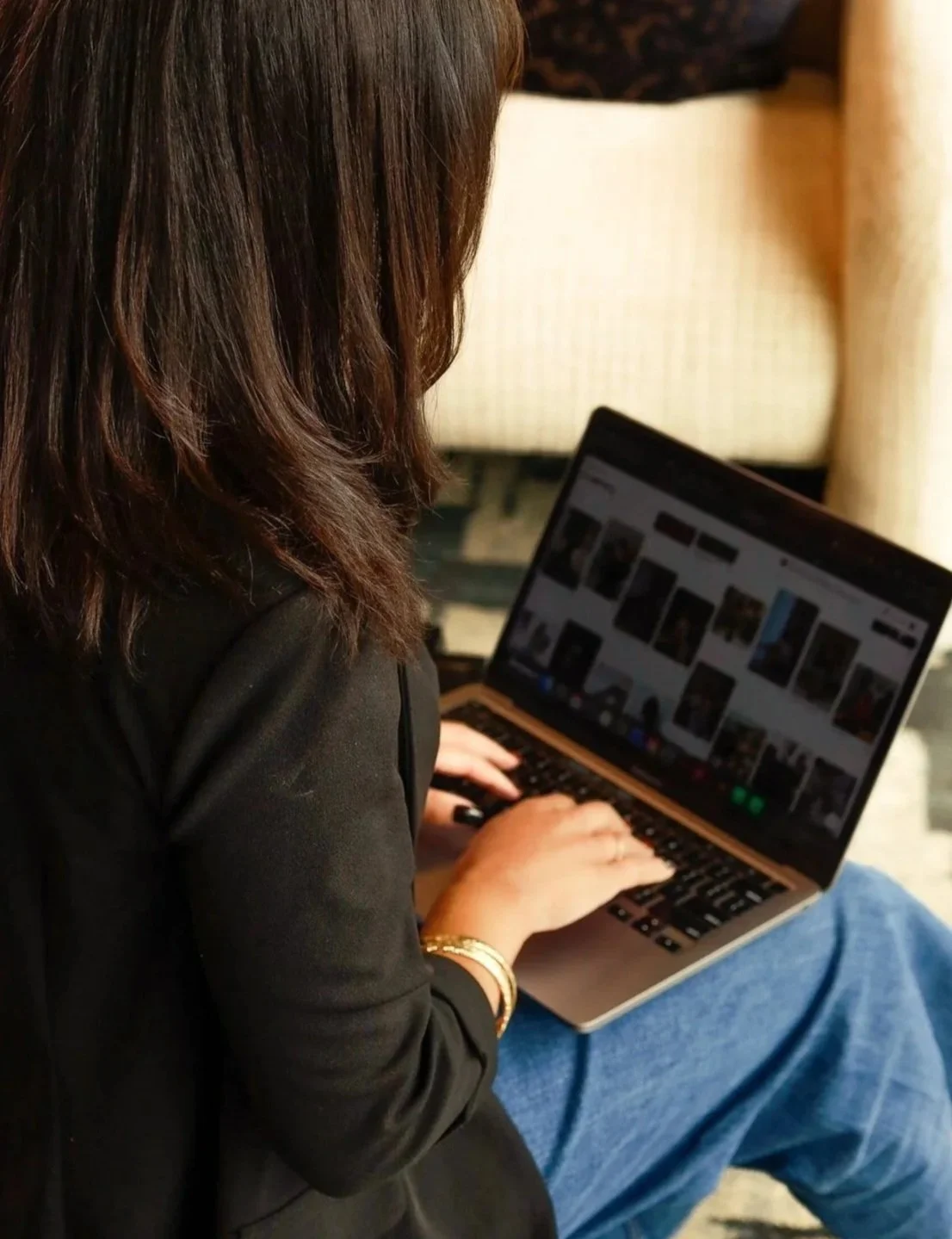Woman with dark hair using a laptop, sitting on a chair with a beige cushion in the background.