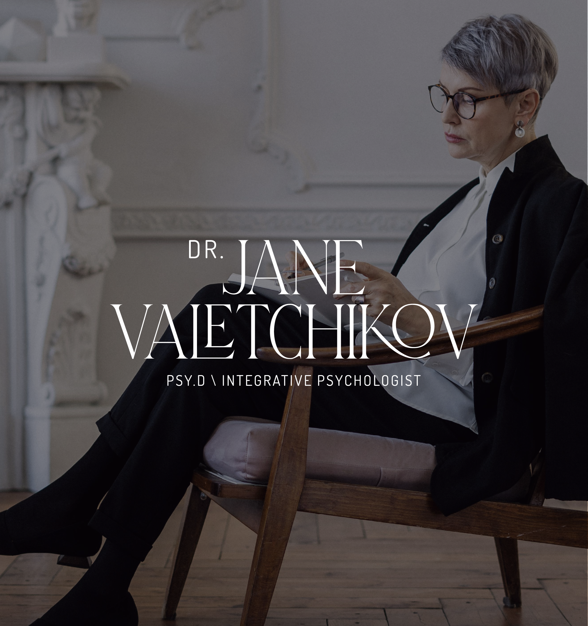 A woman with short gray hair, glasses, and earrings sitting in a wooden chair, reading a book in a room with decorative architectural details.