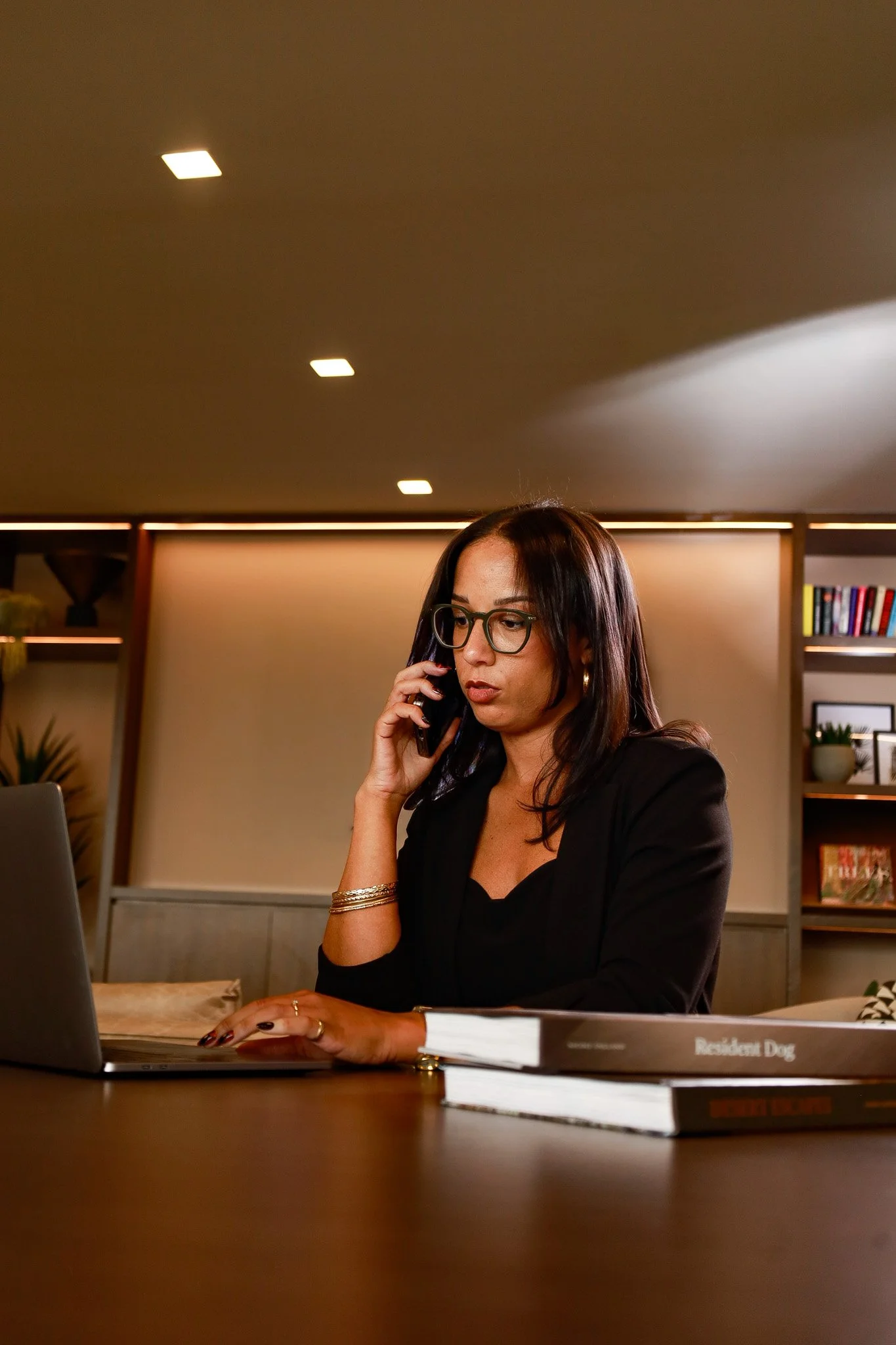 A woman with dark brown hair and glasses working on a laptop, talking on a phone, seated at a desk in an office with bookshelves and decorative items in the background.