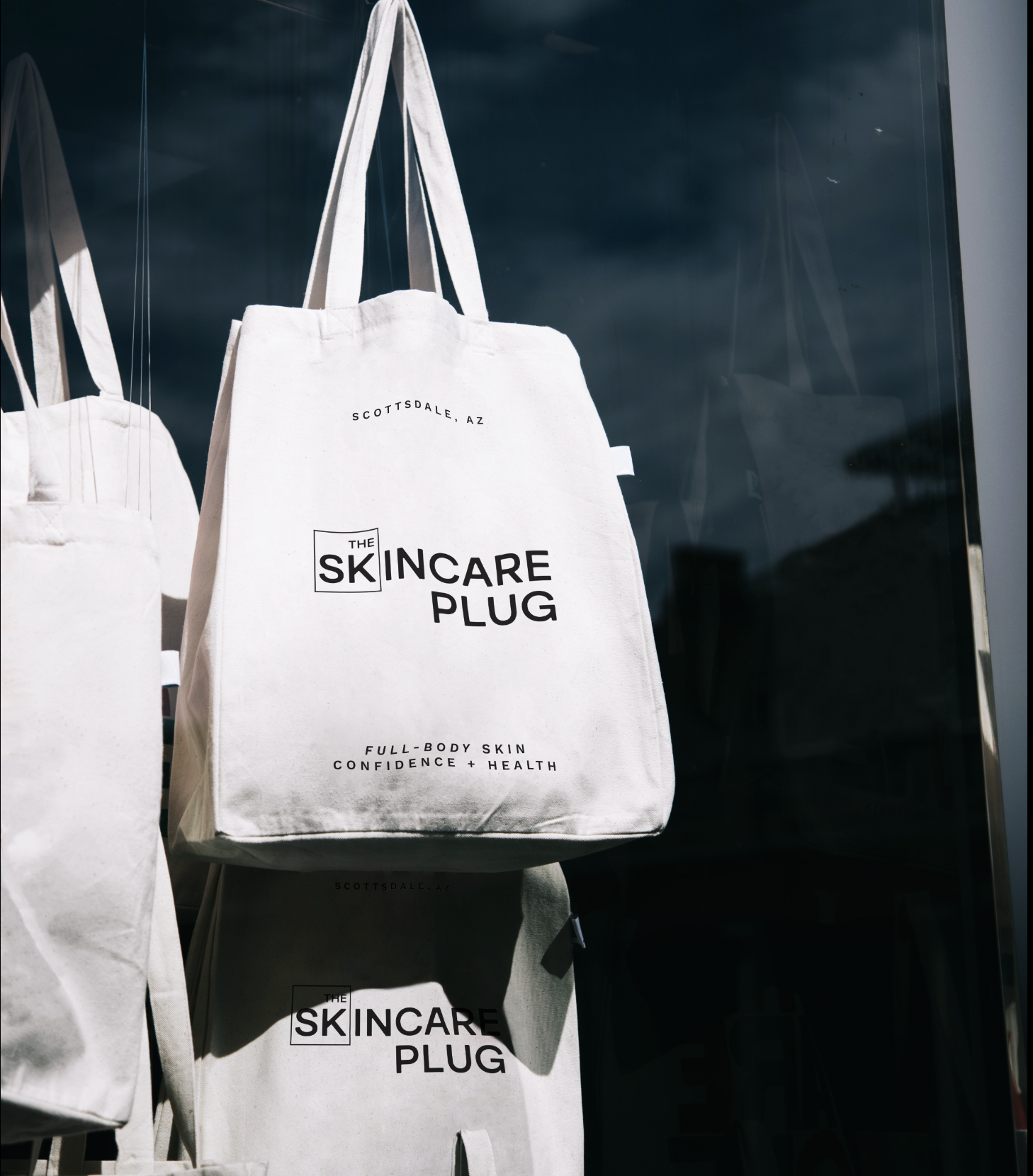 White tote bag with black text reading "The Skincare Plug," hanging on display outdoors, with a reflective surface in the background.