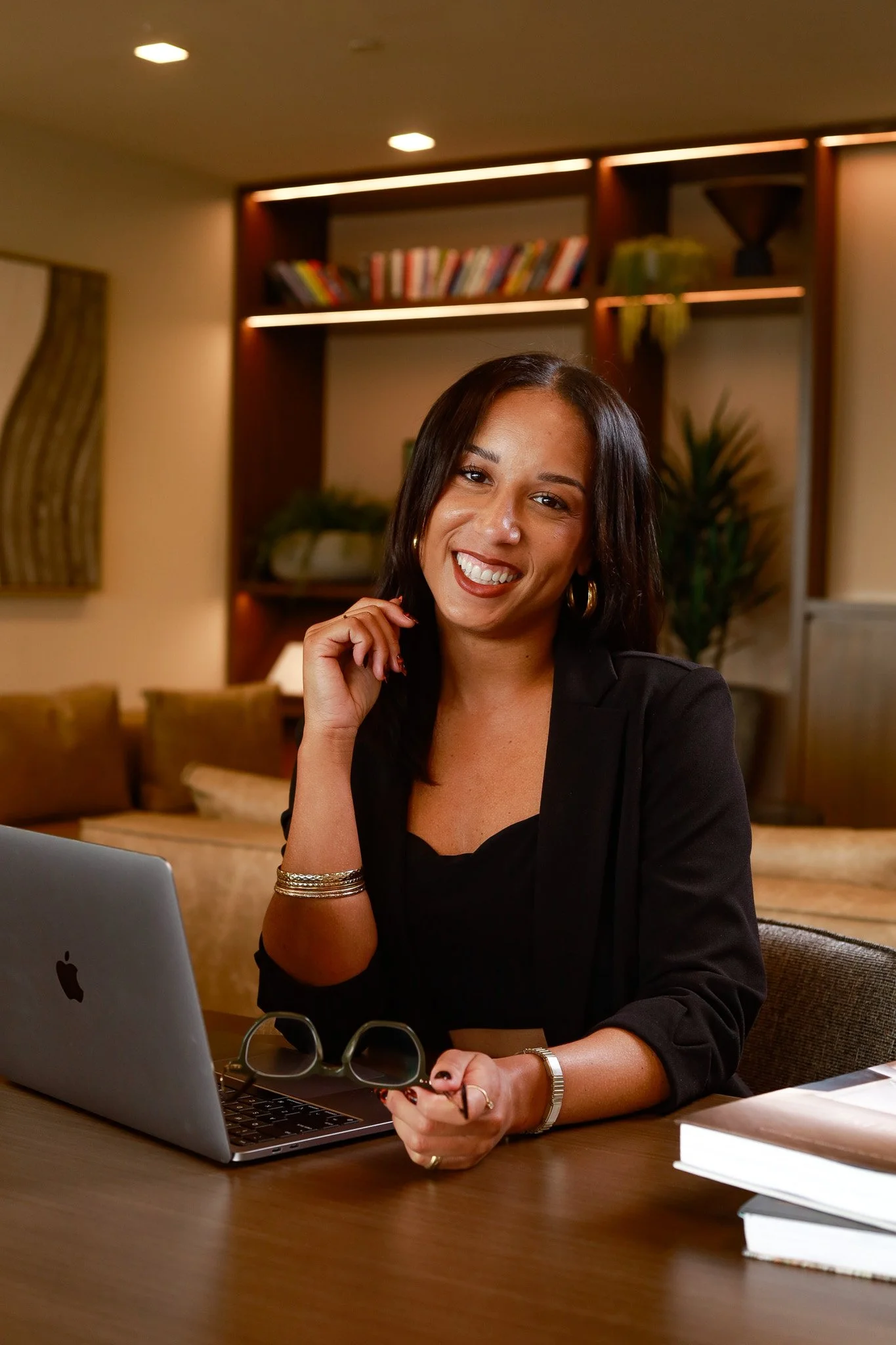 A woman with shoulder-length dark hair smiling, sitting at a desk with a laptop, holding her eyeglasses in her right hand, in a cozy office or living room setting with bookshelves, plants, and warm lighting.