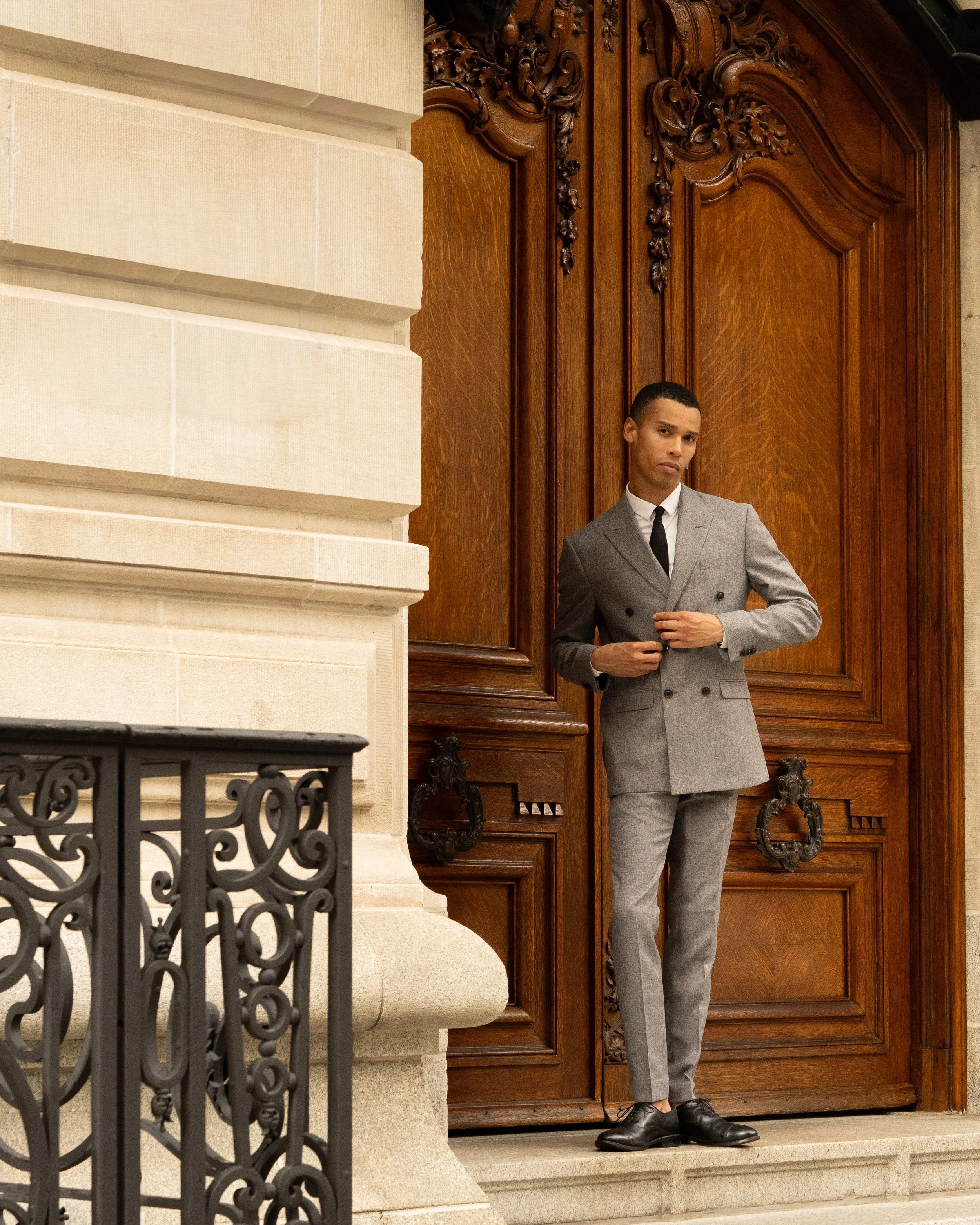 A young man in a gray suit stands on the steps outside a building with large wooden doors and ornate carvings. Part of a stone wall and a decorative black iron railing are visible in the foreground.