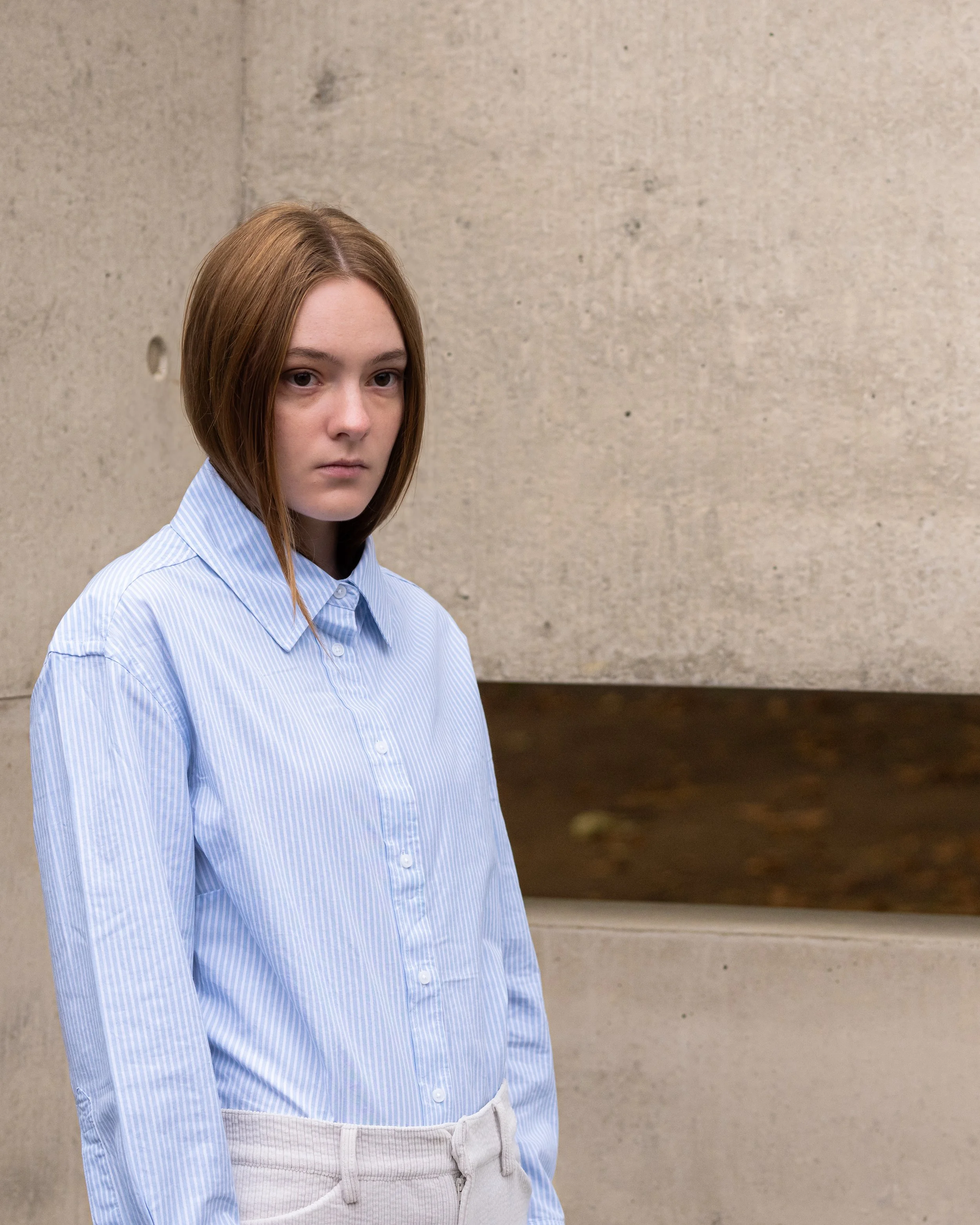 A young woman with straight, shoulder-length brown hair and light skin stands against a beige wall, wearing a light blue and white striped button-up shirt and beige pants.