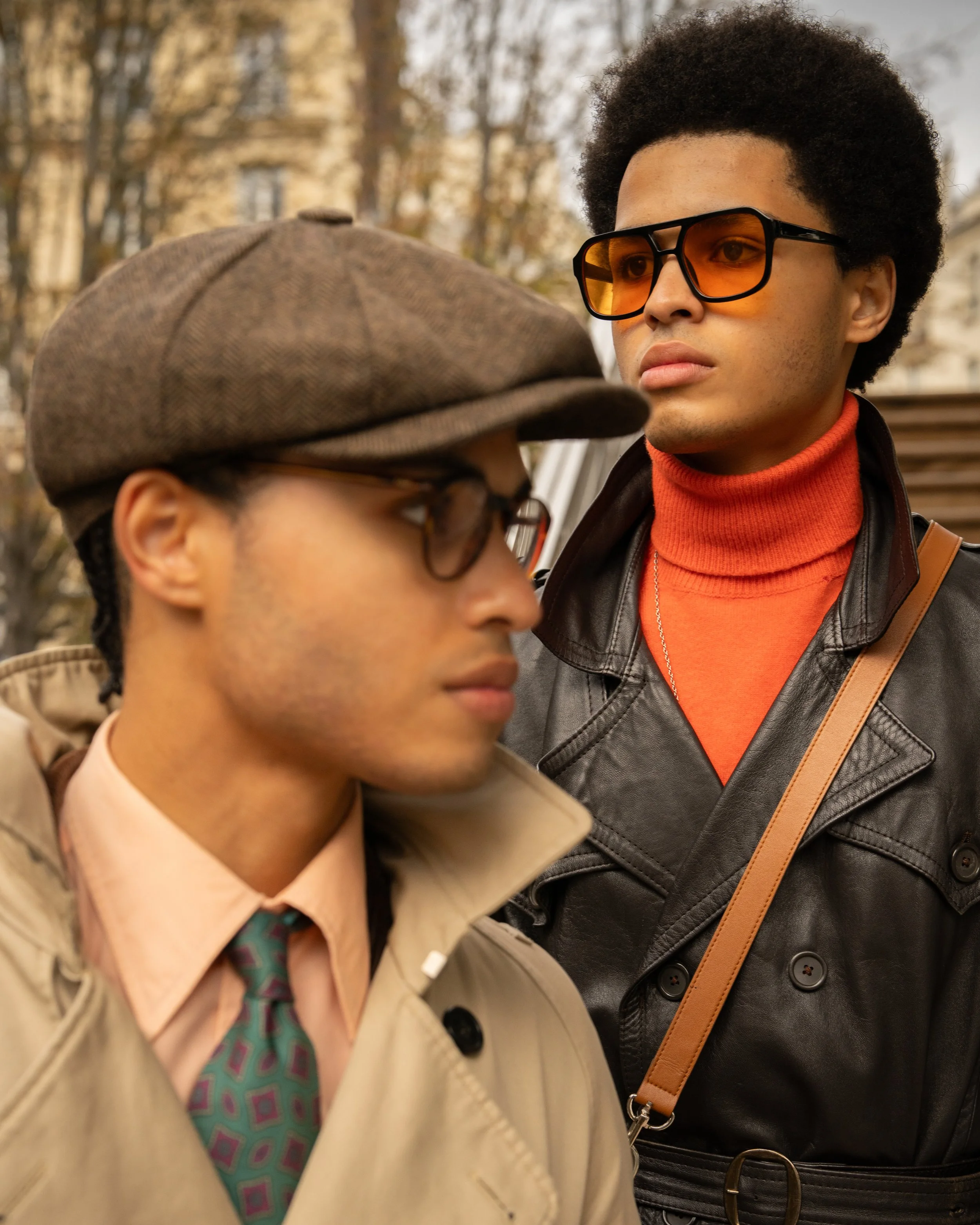 Two young men with dark curly hair face each other outdoors. One is wearing orange-tinted glasses, a black leather jacket, and an orange turtleneck. The other has on a brown cap, beige trench coat, and patterned tie. The background features leafless trees and a neutral building, suggesting autumn.
