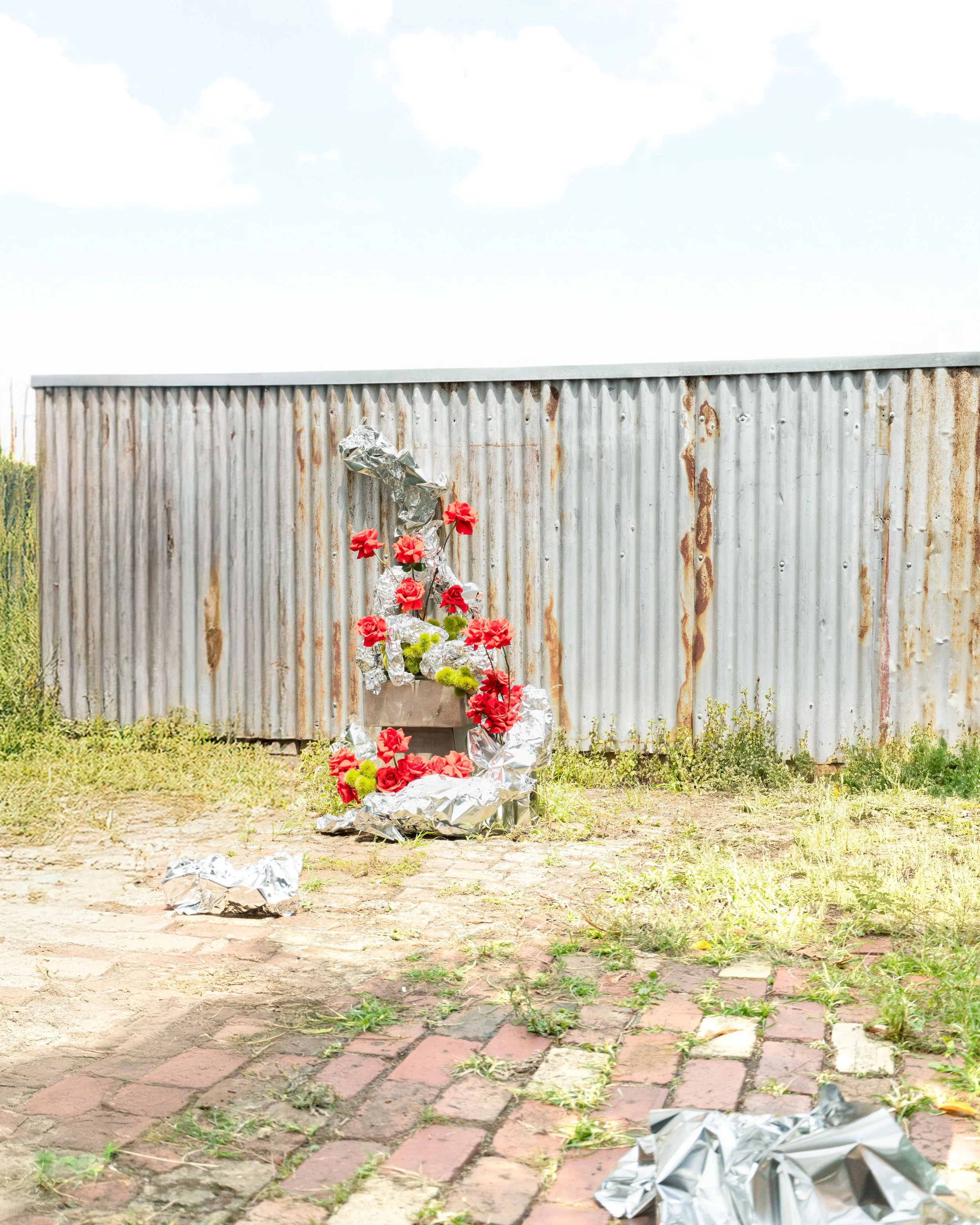 A memorial placed in front of a rusty metal wall with flowers and crumpled foil on the ground.