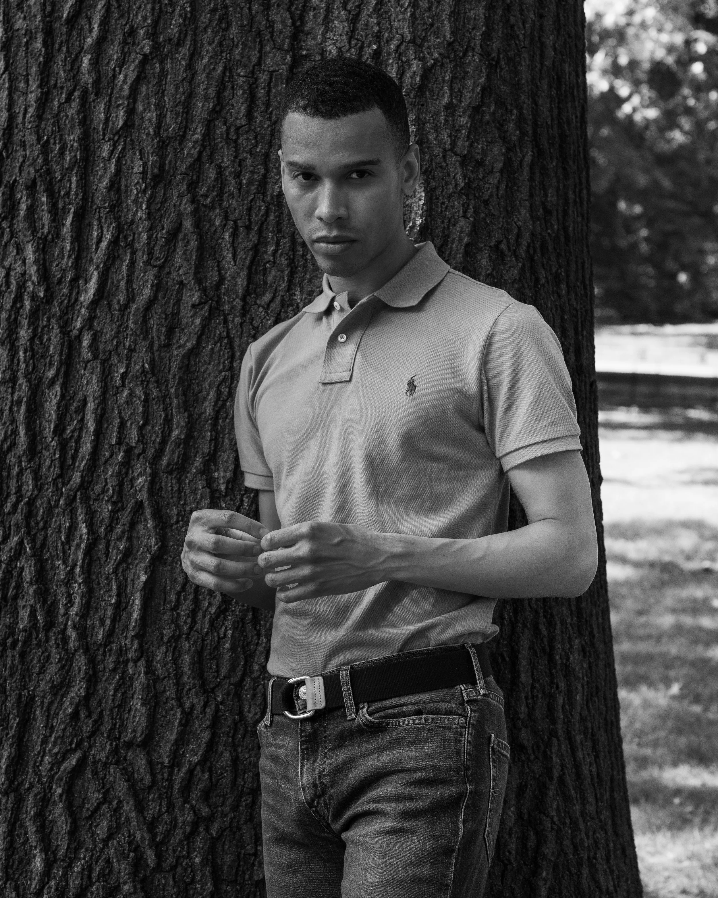 A young man leaning against a tree outdoors, wearing a beige polo shirt and jeans, looking directly at the camera.