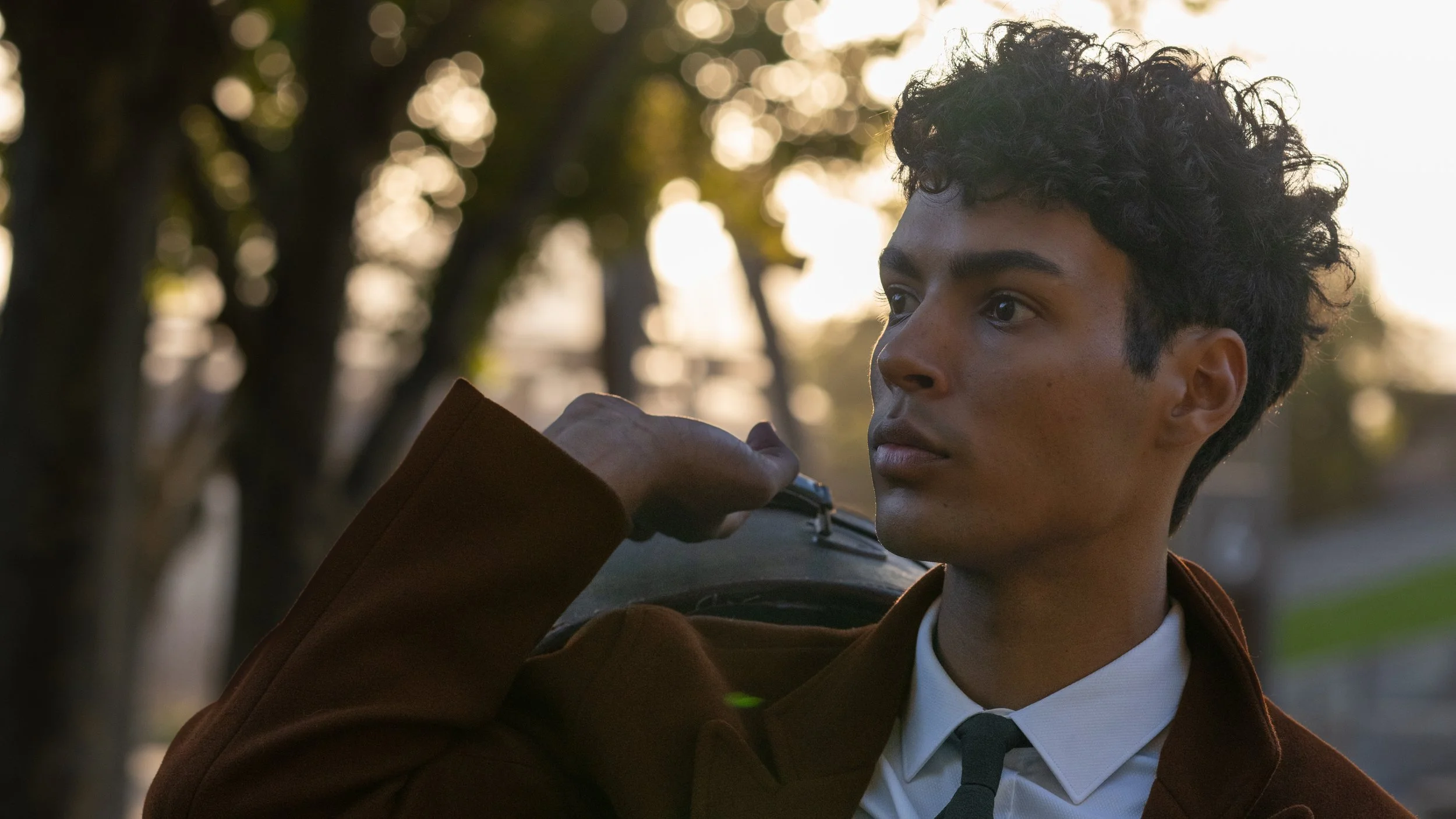Young man with curly dark hair in a brown coat and white shirt, outdoors in the evening, adjusting his tie, with sunlight and trees in the background.