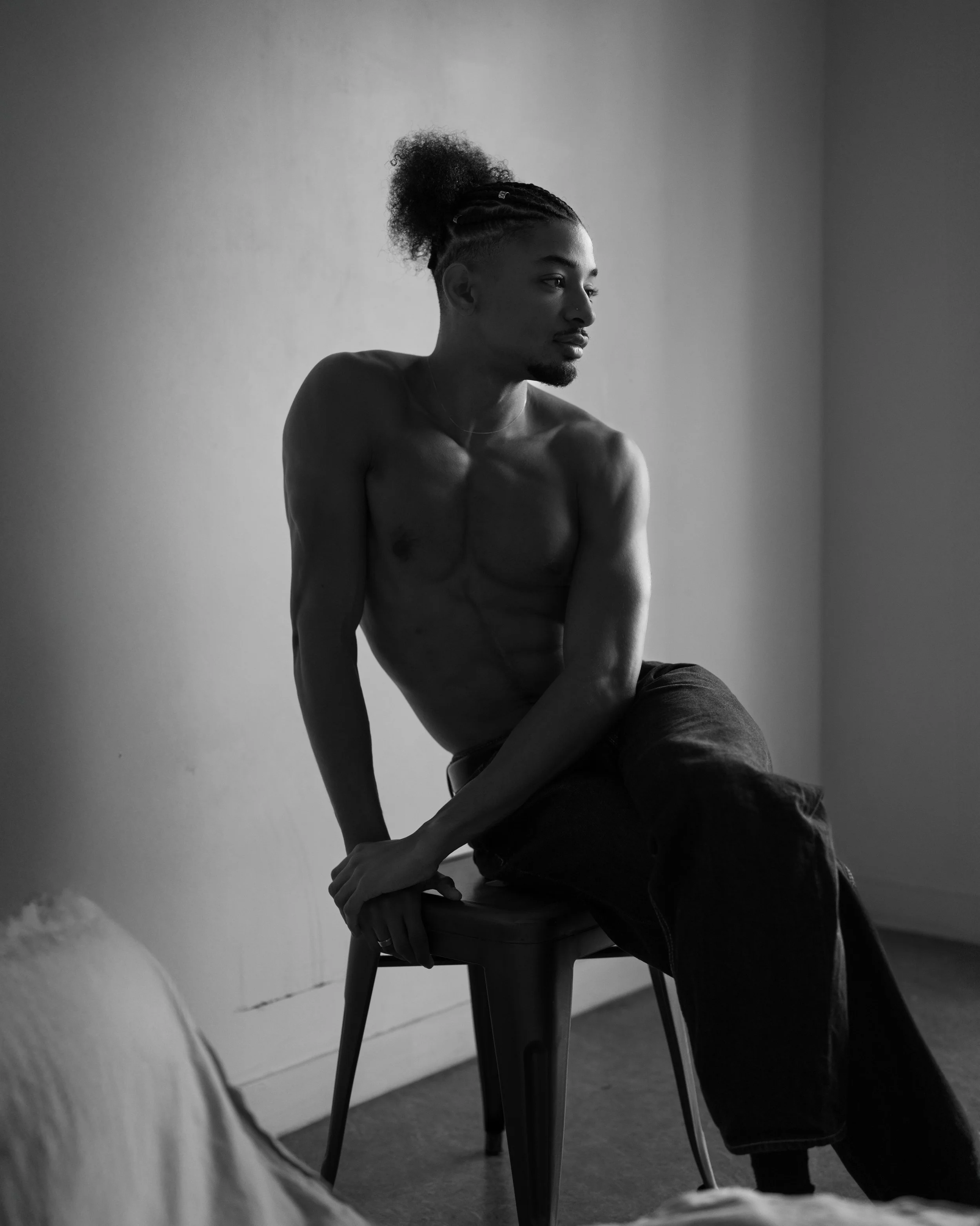 A black-and-white photo of a shirtless young man with styled hair sitting on a chair, with a neutral expression, against a plain wall.