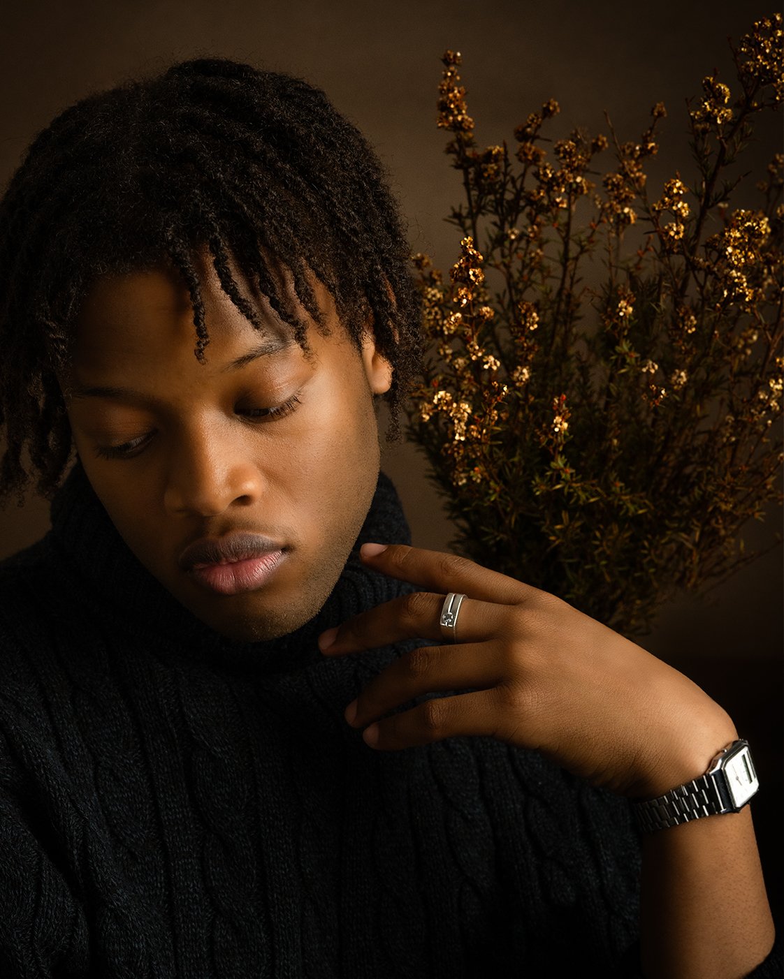 A young man with textured black hair wearing a black sweater, with a silver watch and a ring, looking down with closed eyes against a dark background with a plant.