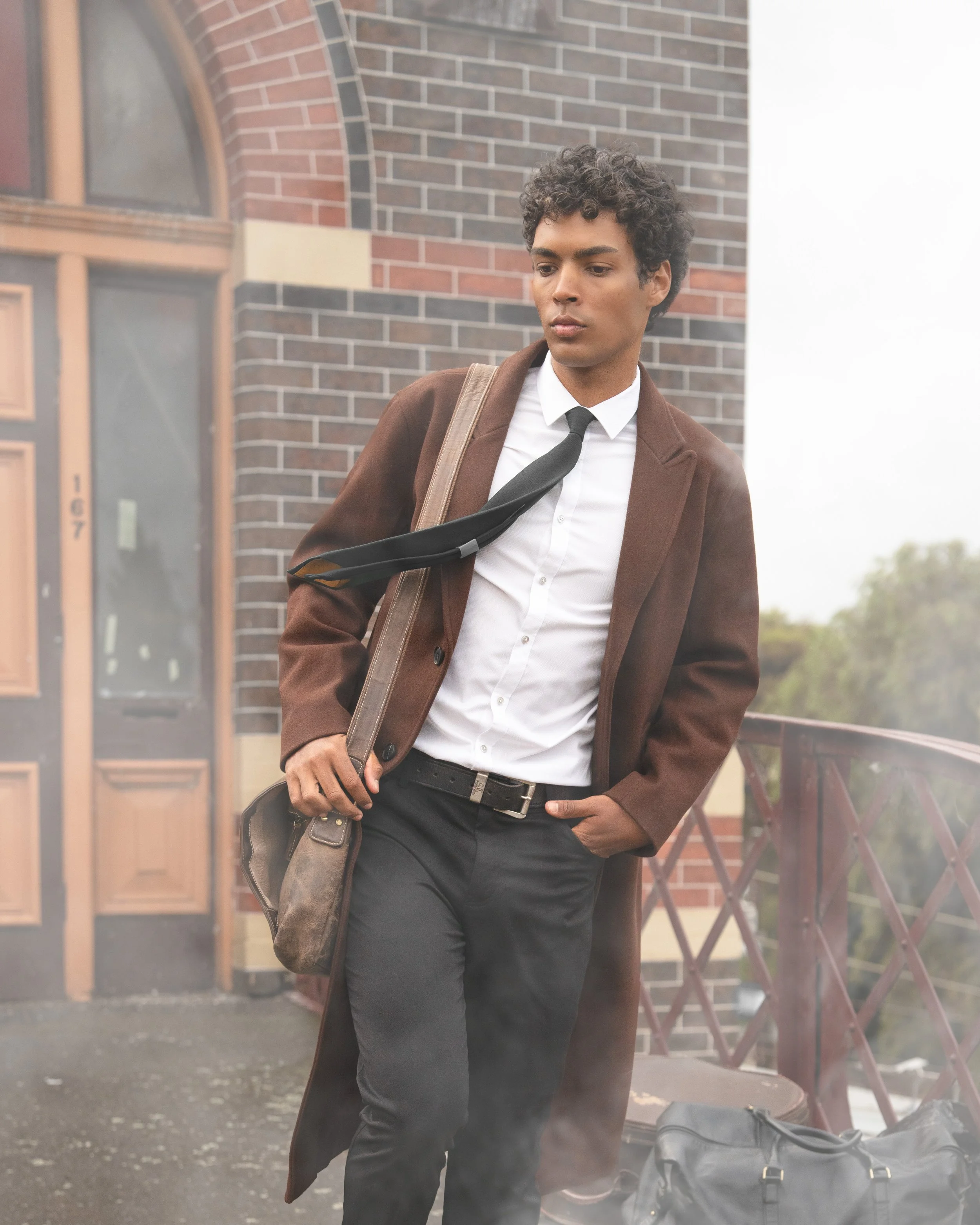 A young man with curly dark hair, wearing a brown coat, white shirt, black tie, and dark pants, stands outdoors near a brick building with an arched window. He carries a tan shoulder bag and has a black duffel bag on the ground nearby, with a foggy or smoky background.