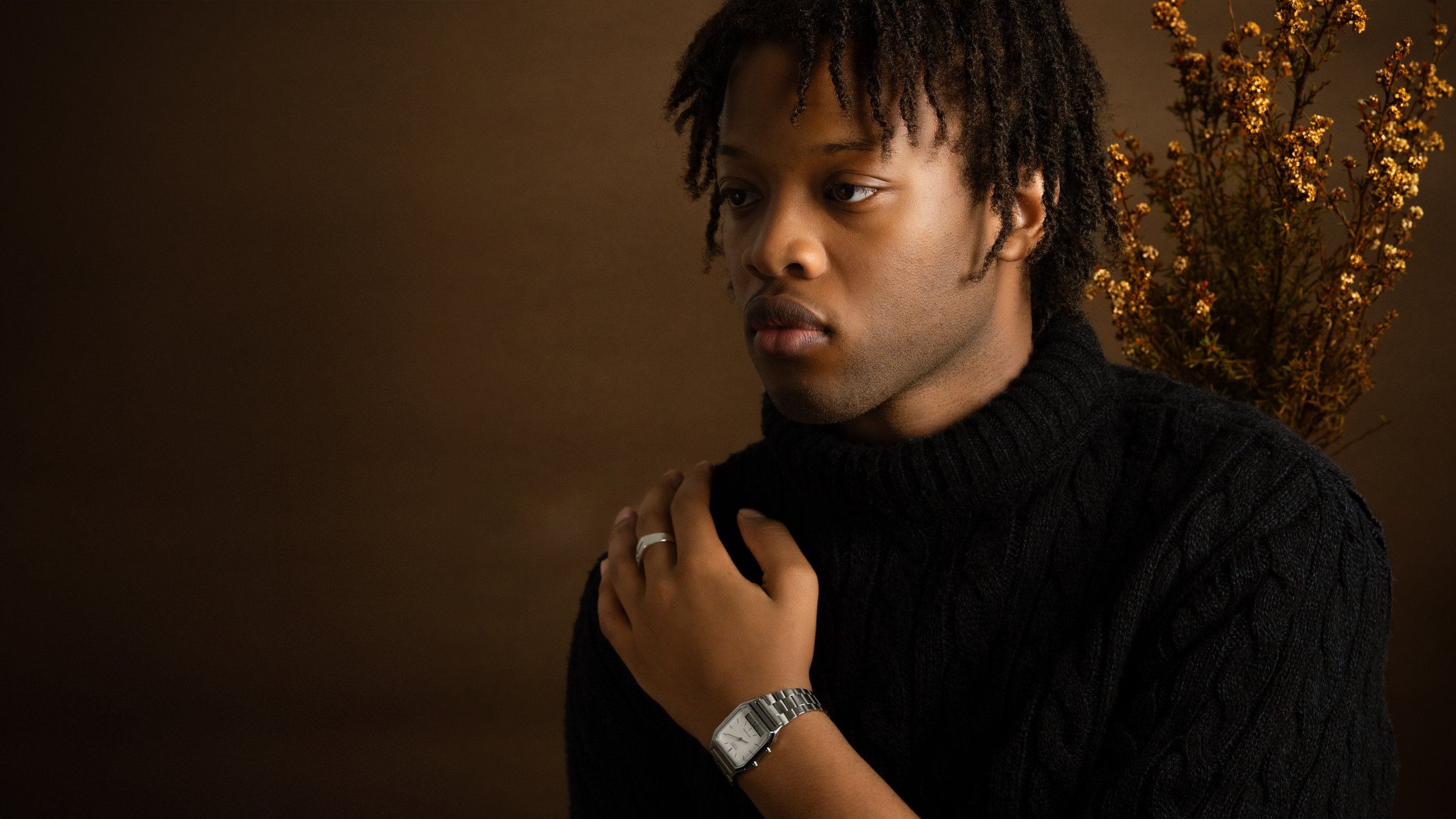A young man with dark skin and dreadlocks wearing a black turtleneck sweater, silver watch, and rings, looking thoughtfully to the side with a hand on his chest, against a brown background with a vase of yellow flowers.