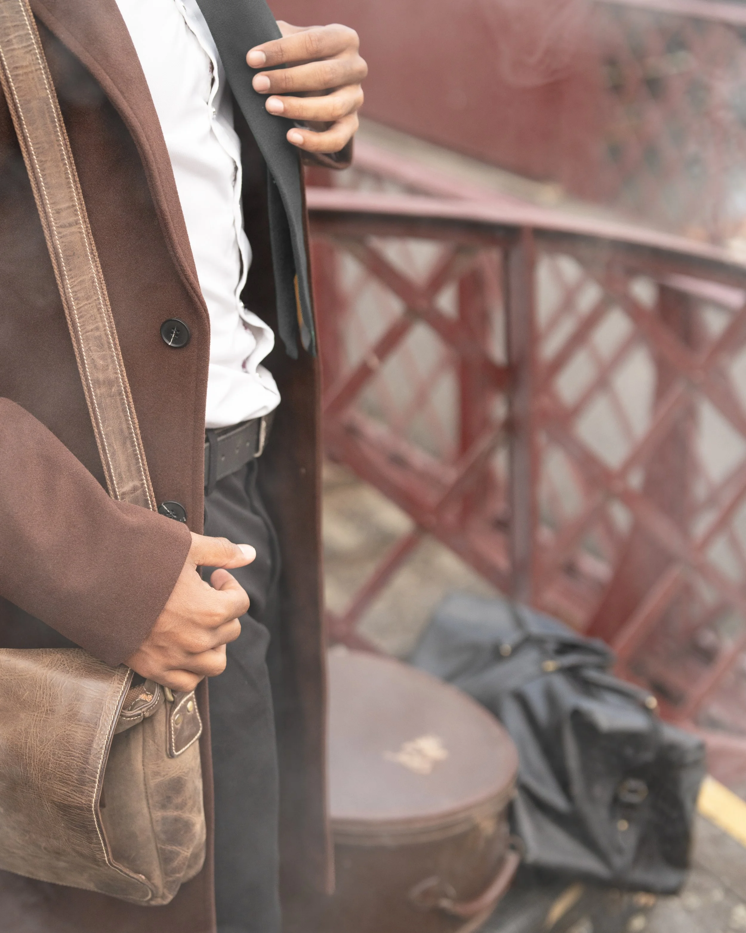 Close-up of two people standing side by side, focusing on their torsos, one wearing a brown jacket and the other in a dark suit, with their hands visible; the person on the left has their hand in their pocket, and the person on the right has their hand resting on their chest. In the background, a red bench and a stone-paved ground are visible.