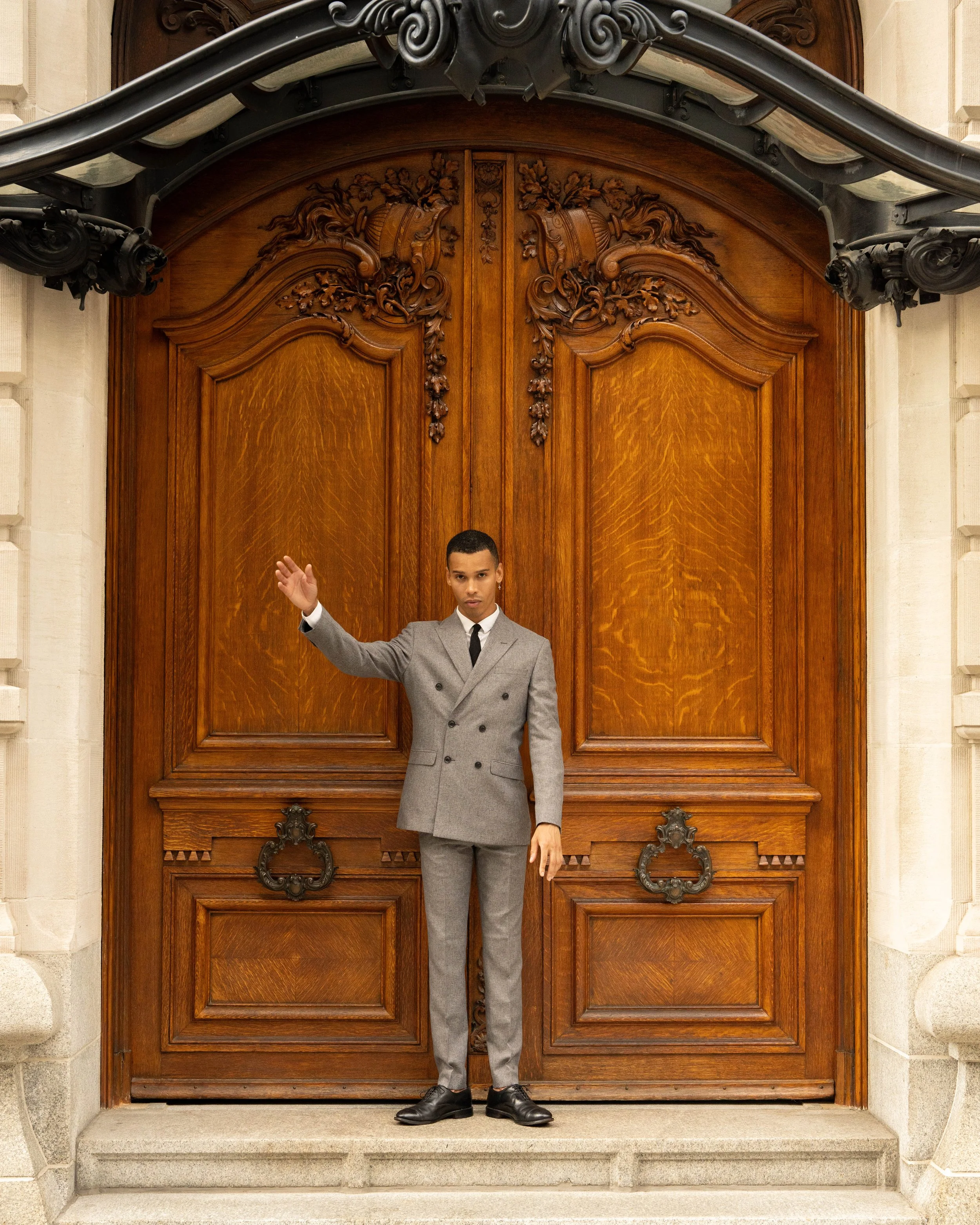 A young man in a gray suit and black shoes standing in front of a large wooden door, raising his right hand with open palm, on a stone step in an ornate building.