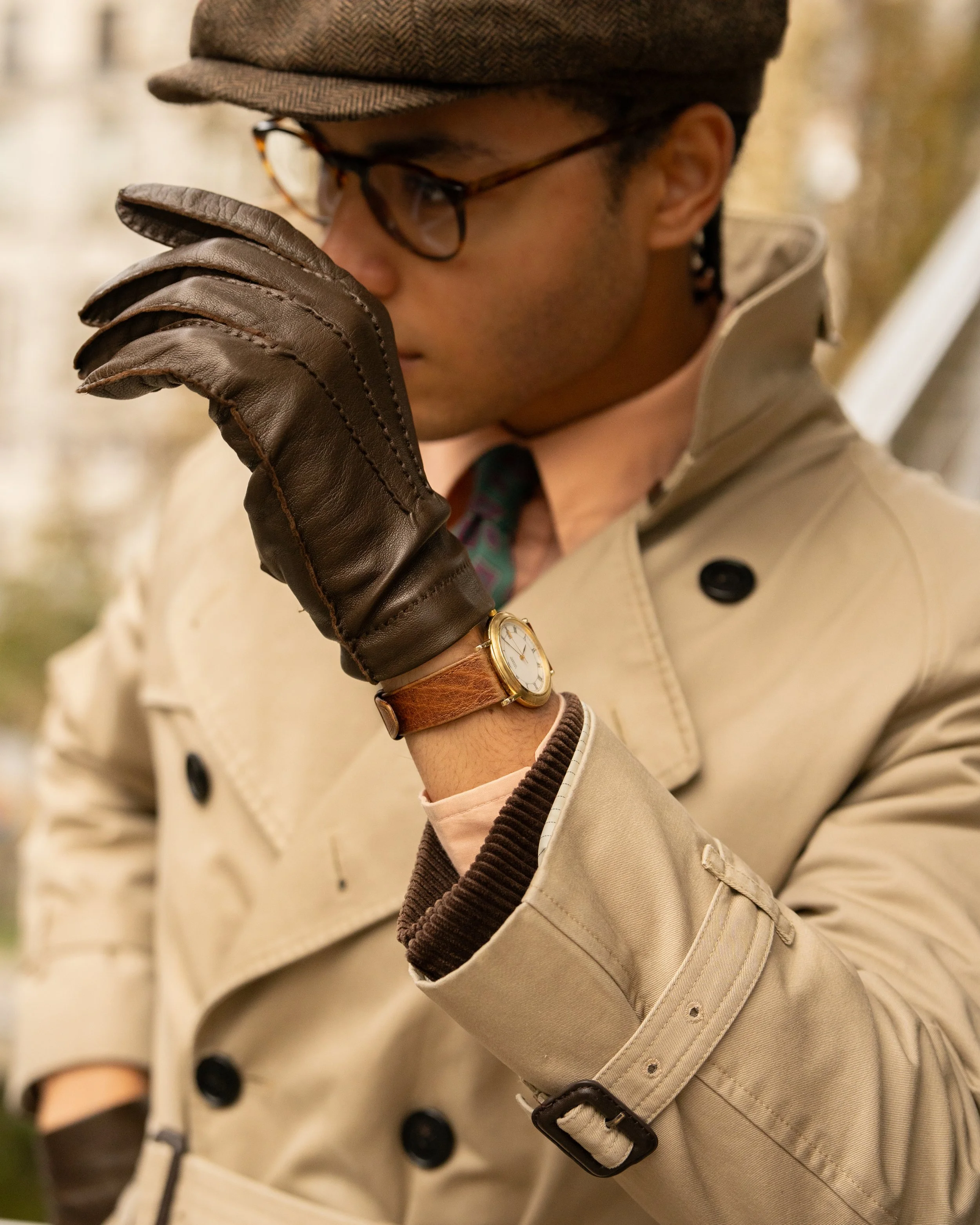 Close-up of a person wearing a brown cap, black glasses, brown leather glove, and a beige trench coat with a belt.