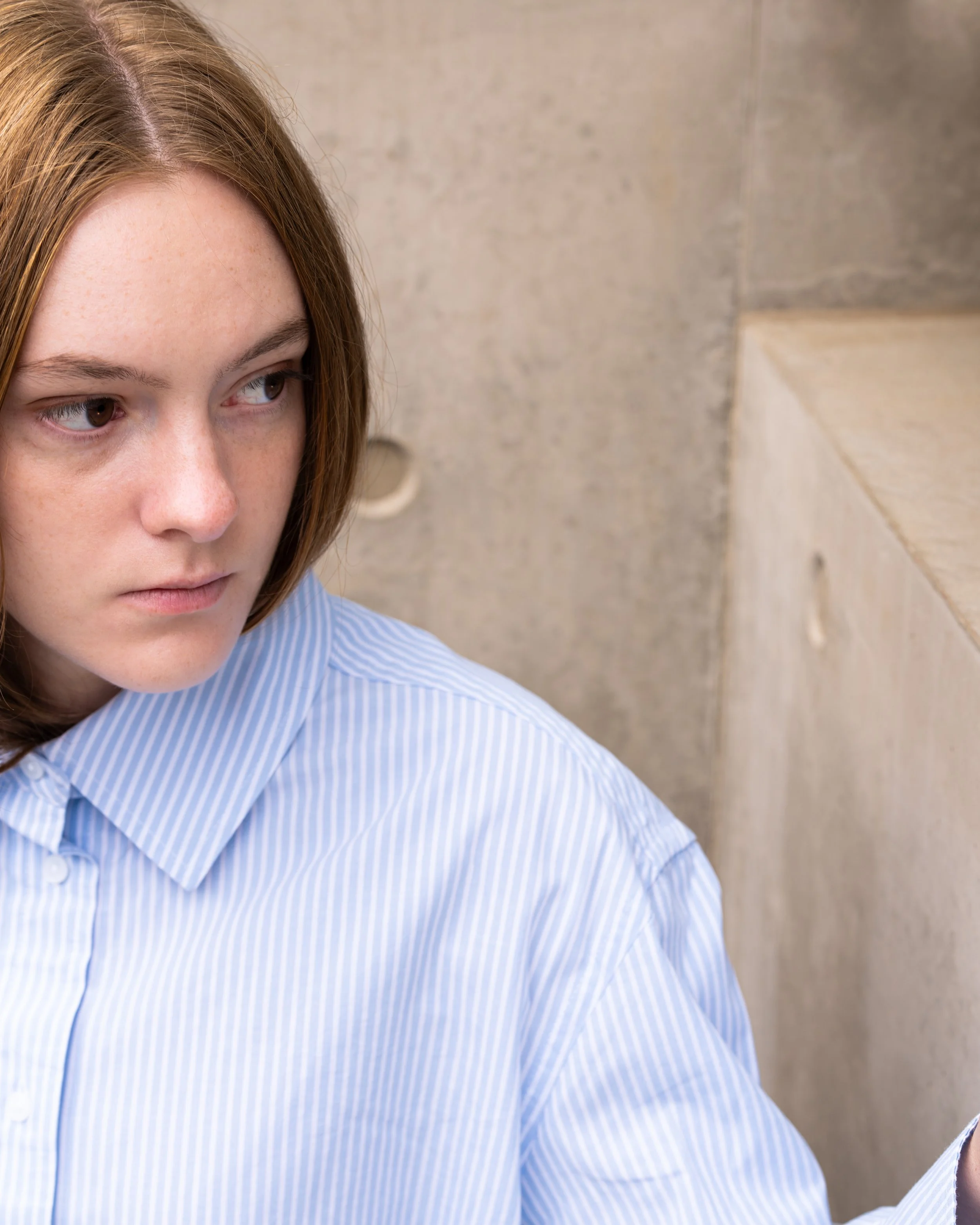 A woman with red hair and freckles wearing a blue striped shirt, sitting against a beige tiled wall.