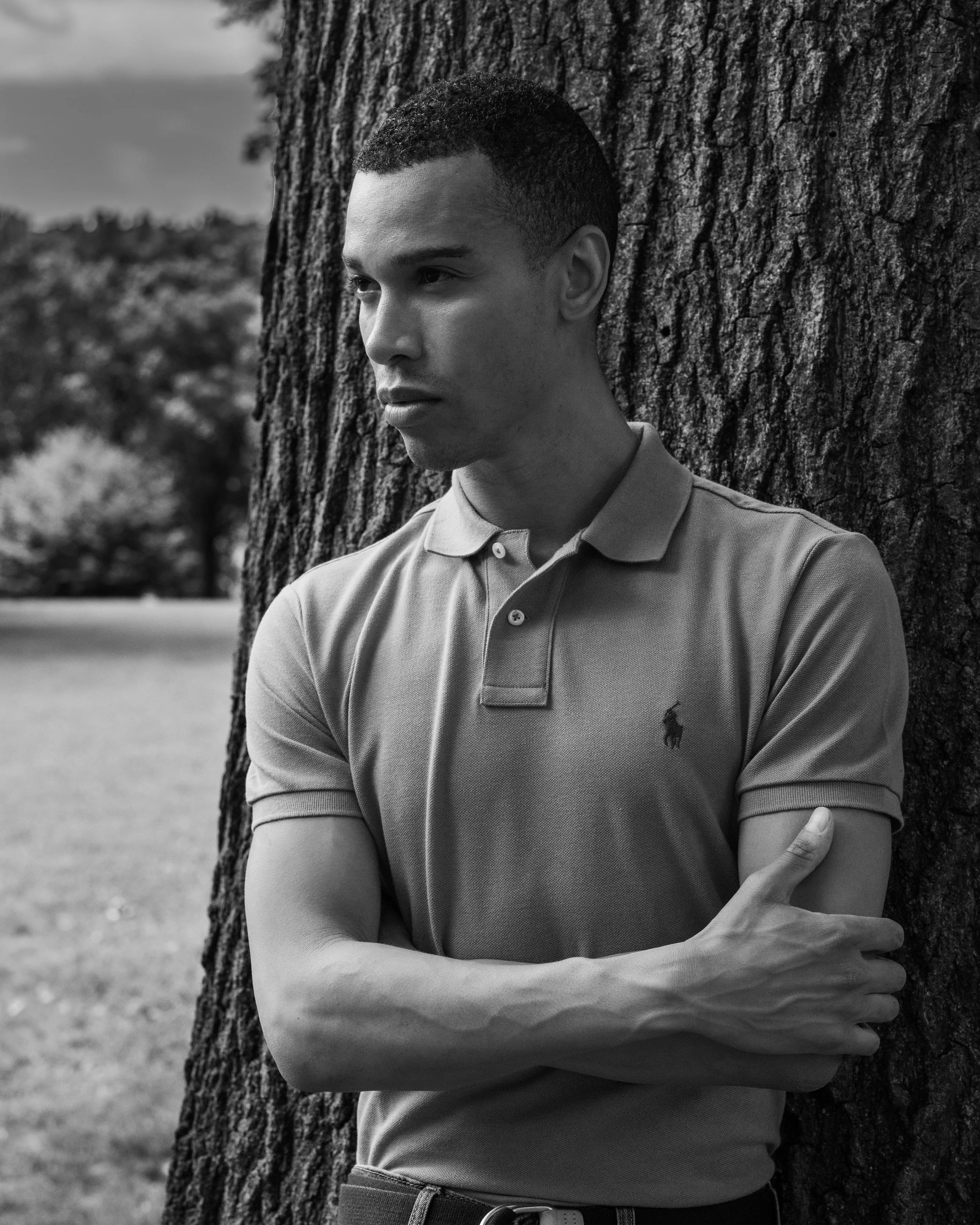 A young man wearing a polo shirt is standing outdoors next to a large tree, with his arms crossed. The background shows a blurred natural landscape. The photo is in black and white.