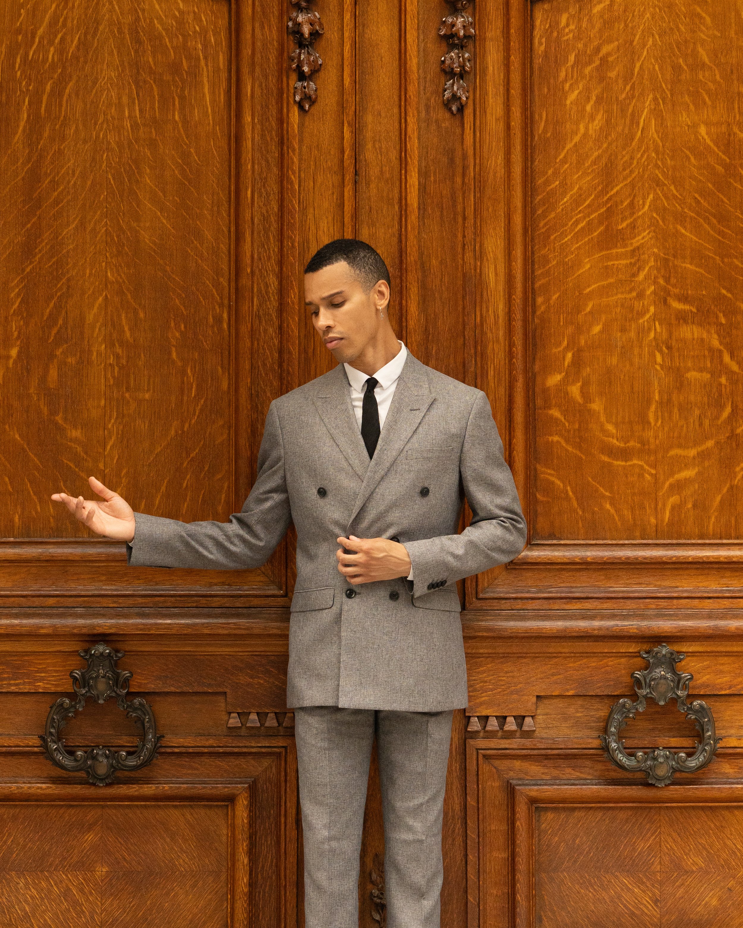 A man dressed in a gray suit with a white shirt and black tie is standing against a large wooden cabinet. The man has short black hair, a light facial hair, and an earring in his left ear. He is looking down with a contemplative expression, with his left hand slightly raised and his right hand resting on his stomach. The wooden cabinet behind him has intricate carved details and a polished finish.