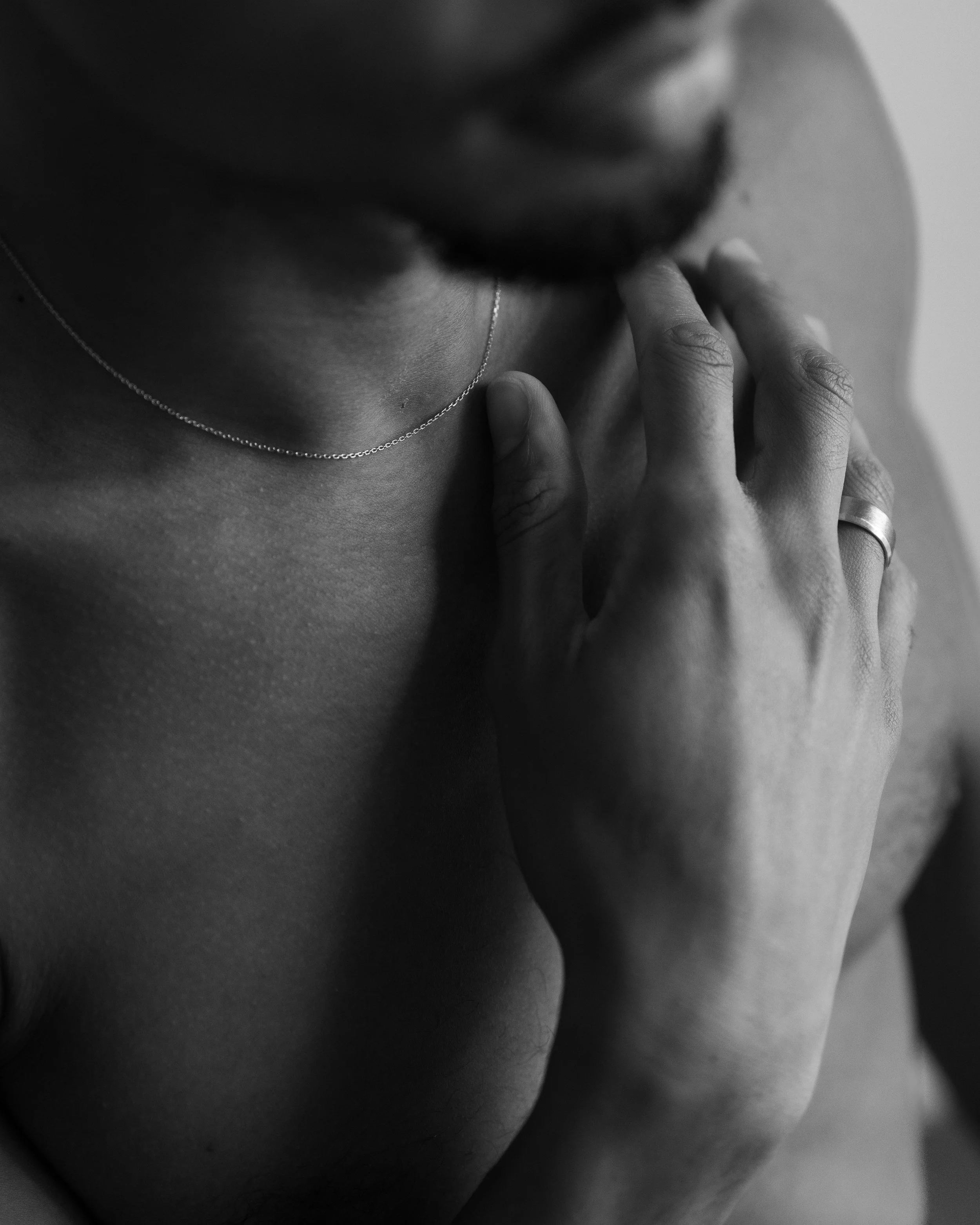 A close-up black and white photograph of a person's face, focusing on their lips, chin, and a hand gently touching the lips. The person is wearing a delicate chain necklace.