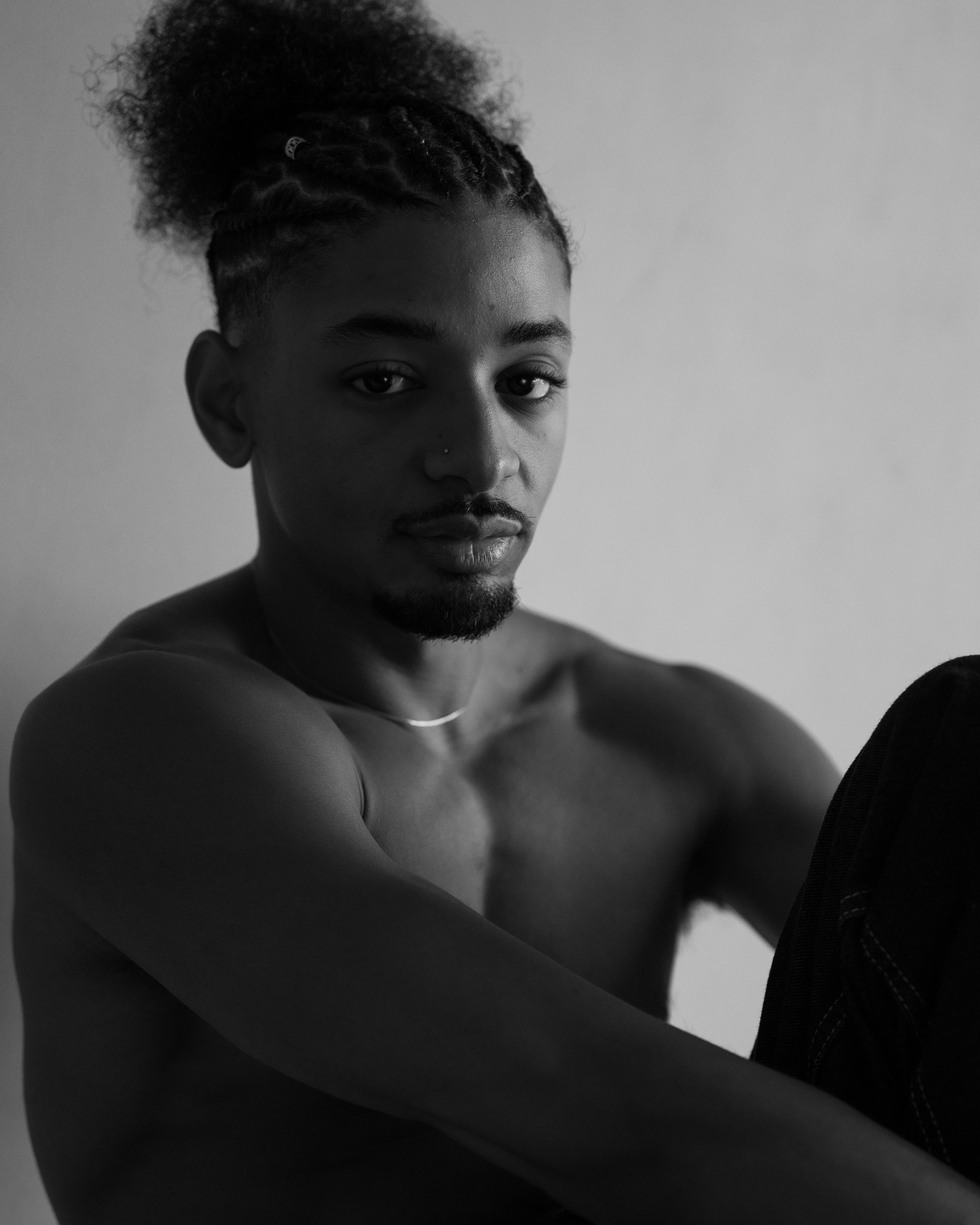 Black and white portrait of a person with short curly hair, some of it braided, posing against a plain wall background.