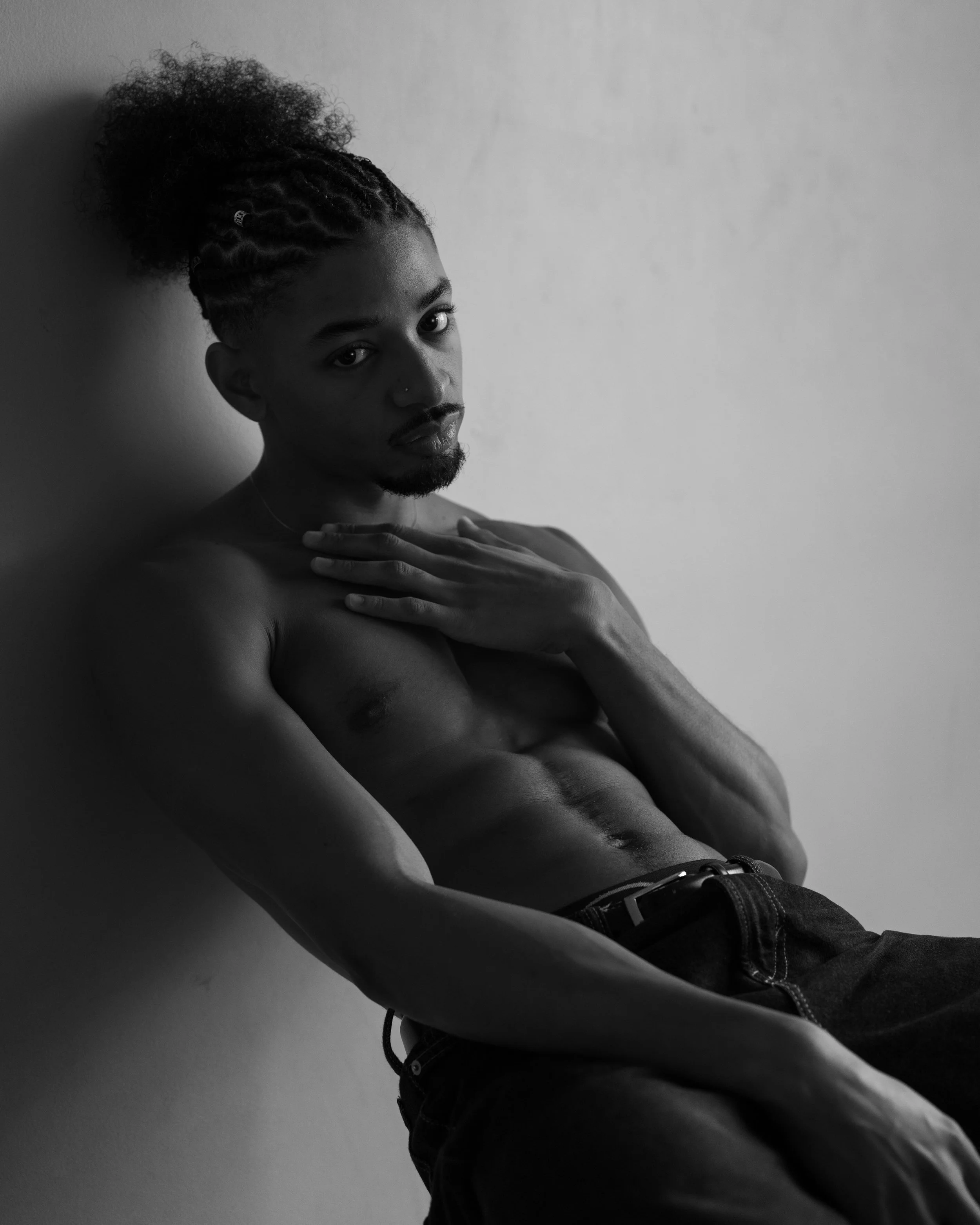 Black and white photo of a shirtless man with curly hair styled in a high curly bun, looking directly at the camera with one hand on his chest, wearing jeans and a belt, against a plain wall background.