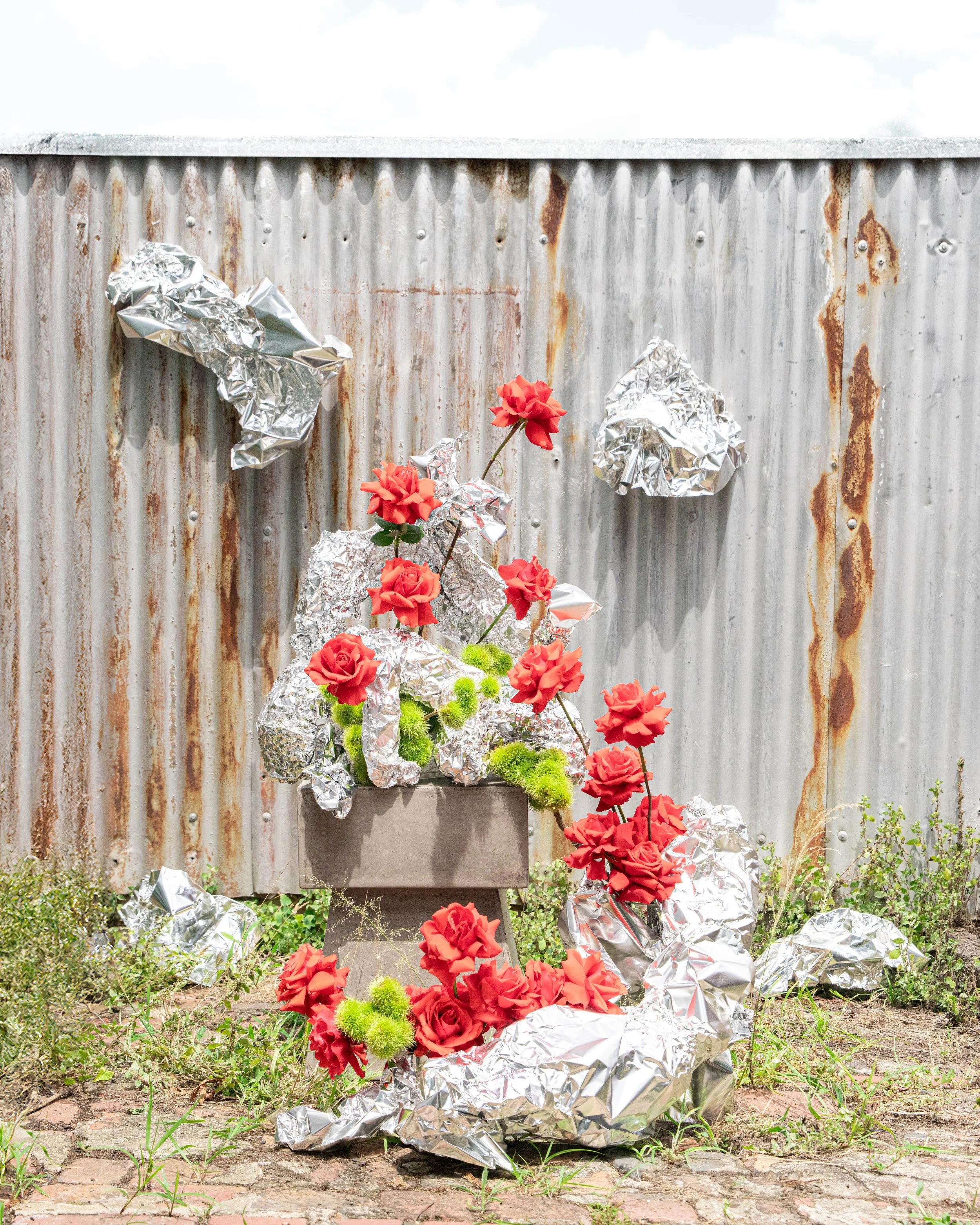 A memorial arrangement of red roses, green chrysanthemums, and aluminum foil on a concrete platform in front of a rusted corrugated metal wall, with scattered flowers and foil at the base on uneven brick ground.