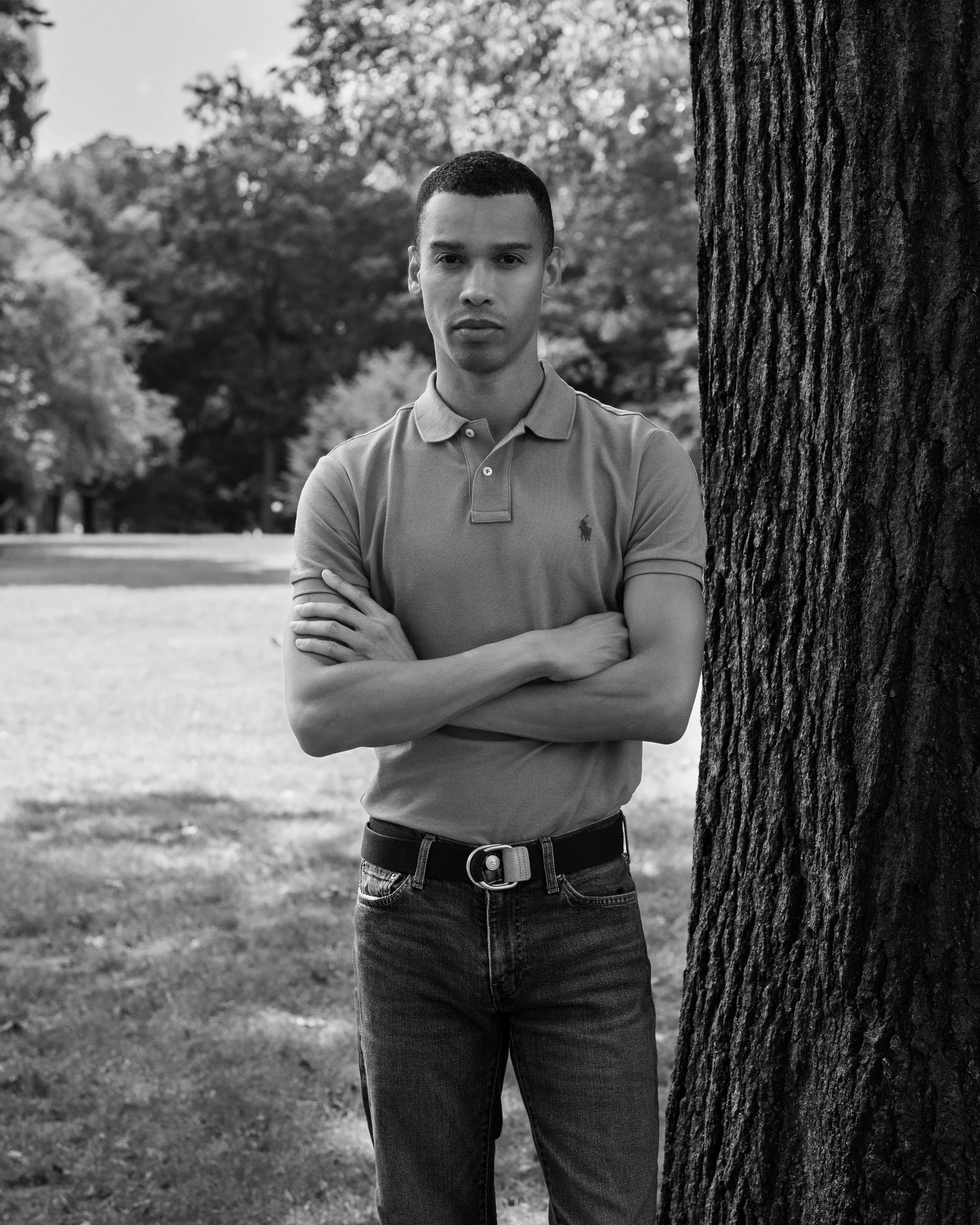 A young man standing outdoors next to a tree with arms crossed. He is wearing a polo shirt and jeans, and gazing toward the camera. The background features blurred trees and a park-like setting in black and white.