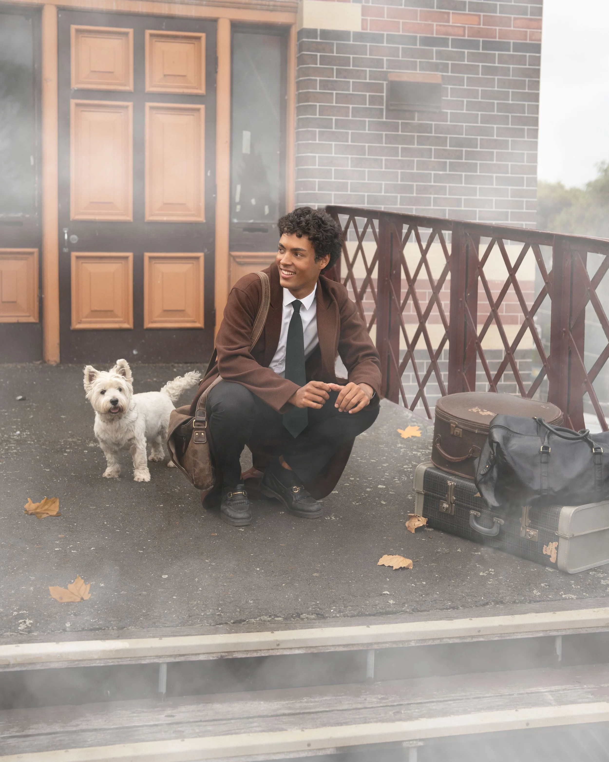 A man in a brown coat and black tie is crouching on a dark pavement outside a brick building, smiling. A white dog with fluffy fur is nearby. There are a few fallen leaves on the ground. Beside the man are a couple of bags and a small black speaker.
